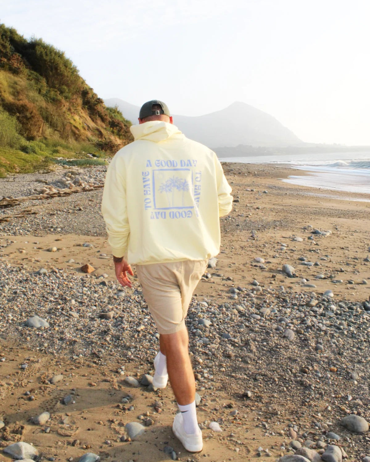 Rückenansicht eines Spaziergangs in butterfarbenem Hoodie entlang eines sonnigen Strandes.
