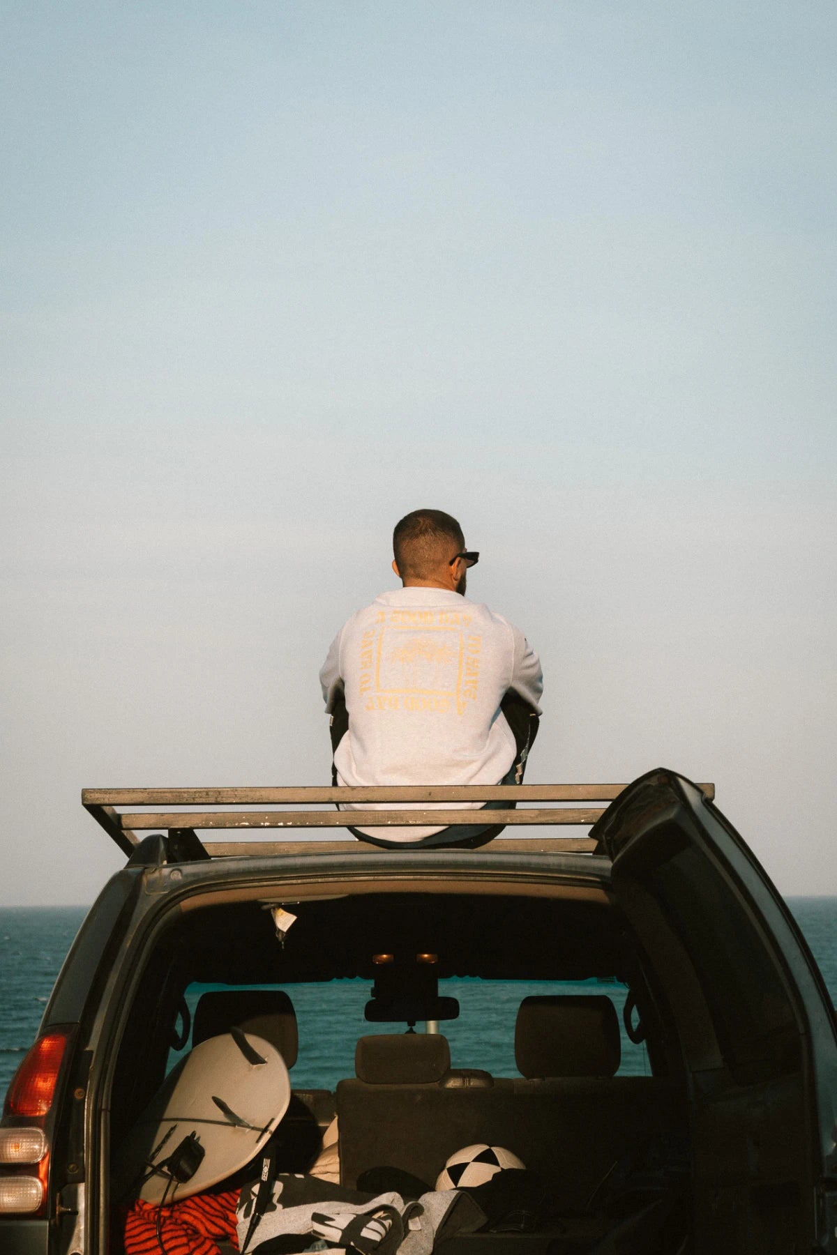 Mann mit dem A Good Day Sweater Cool Grey von The Sunkissed Rebels sitzt auf dem Autodach mit Blick auf den Ozean.