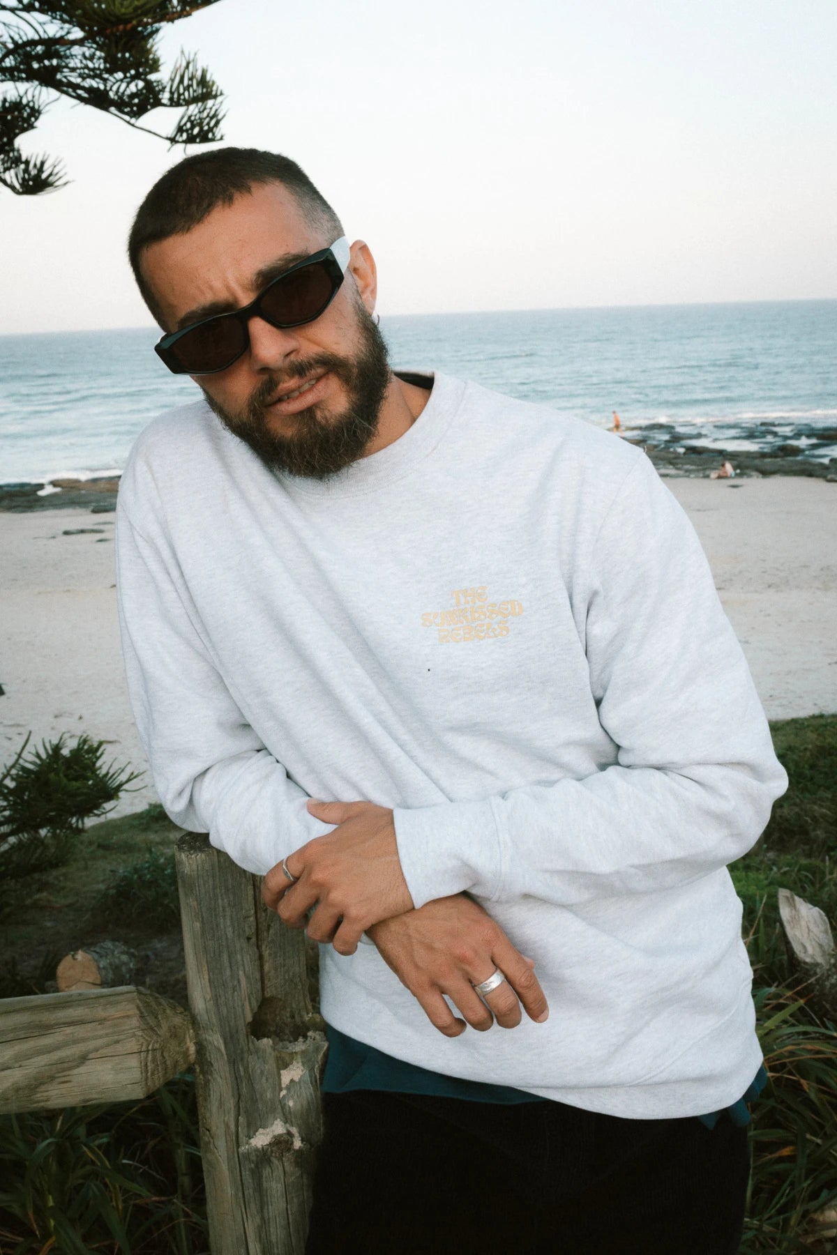 Nahaufnahme – A Good Day Sweater in Grau von The Sunkissed Rebels, getragen von einem Mann mit Sonnenbrille am Meer.