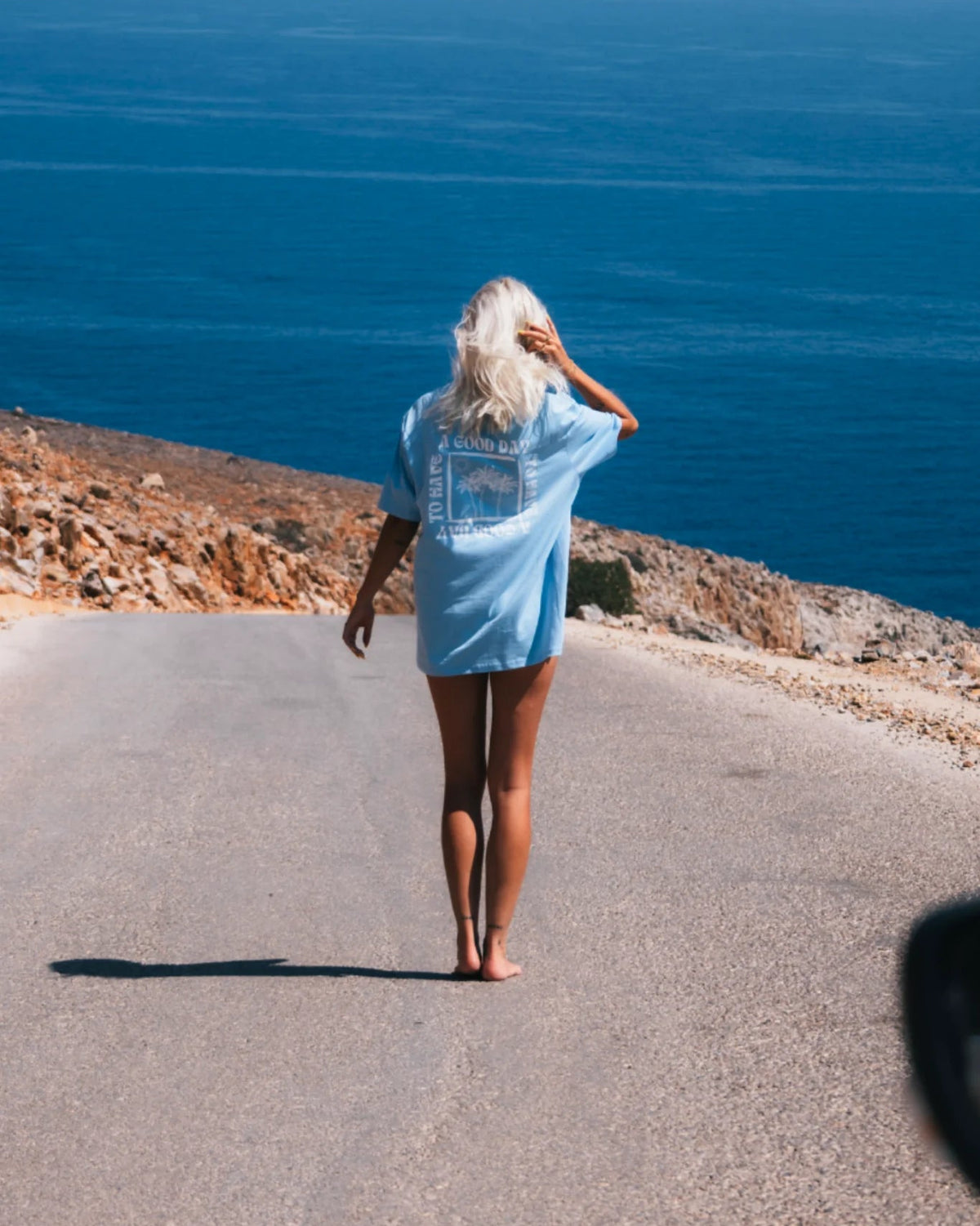 Frau läuft barfuß an Küstenstraße mit blauem A Good Day T-Shirt – Sunkissed Rebels Blue Soul Shirt am Meer