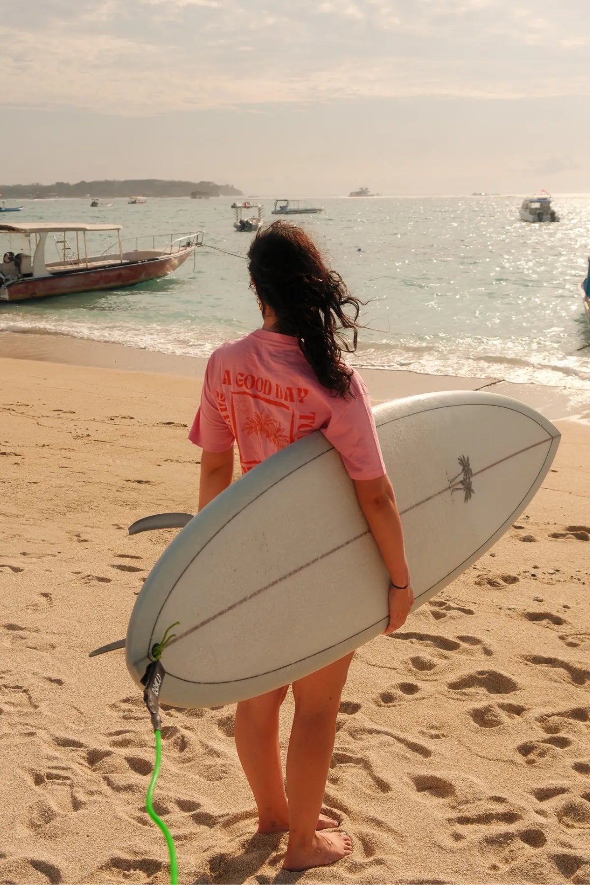 Frau mit Surfbrett am Strand trägt das A Good Day Women T-Shirt Pink von The Sunkissed Rebels – sommerliche Beach-Vibes.