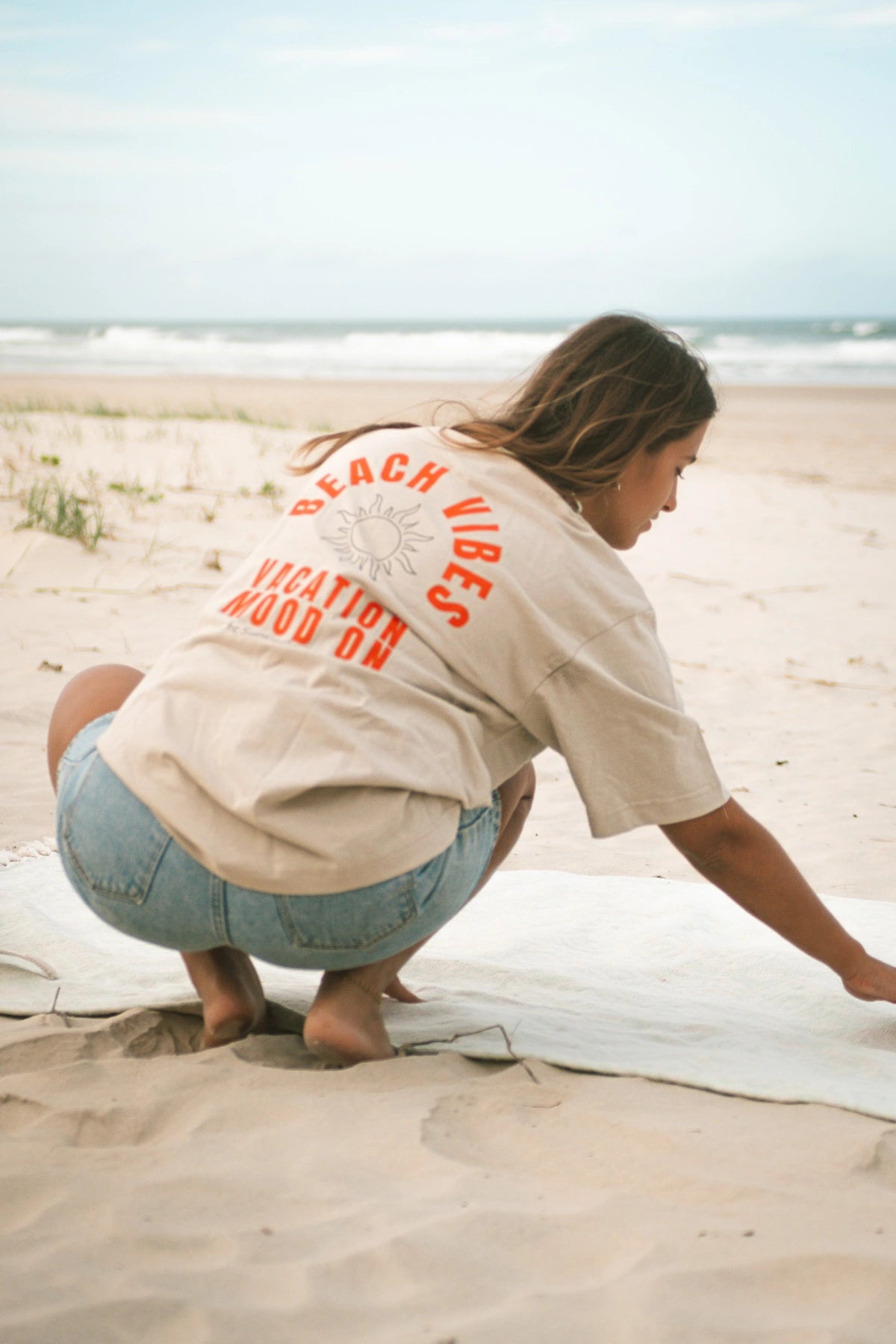 Frau richtet ihre Decke am Strand – Fokus auf Rückenaufdruck Beach Vibes Oversized T-Shirt in Sand