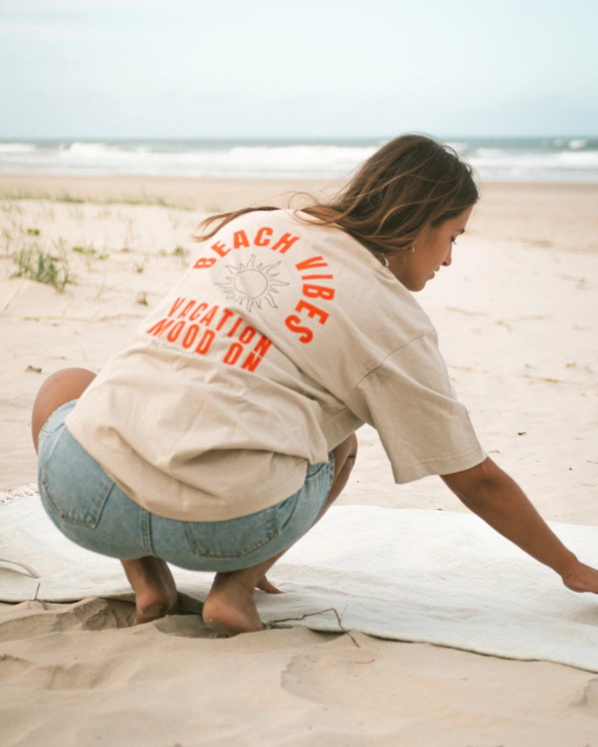 Frau richtet ihre Decke am Strand – Fokus auf Rückenaufdruck Beach Vibes Oversized T-Shirt in Sand