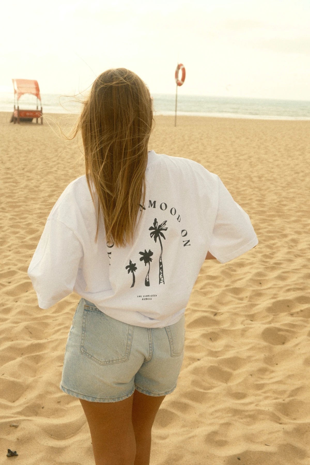Back view of a woman at sunset in the sand wearing the Vacation Mood On T-shirt – ocean horizon and lifeguard vibes