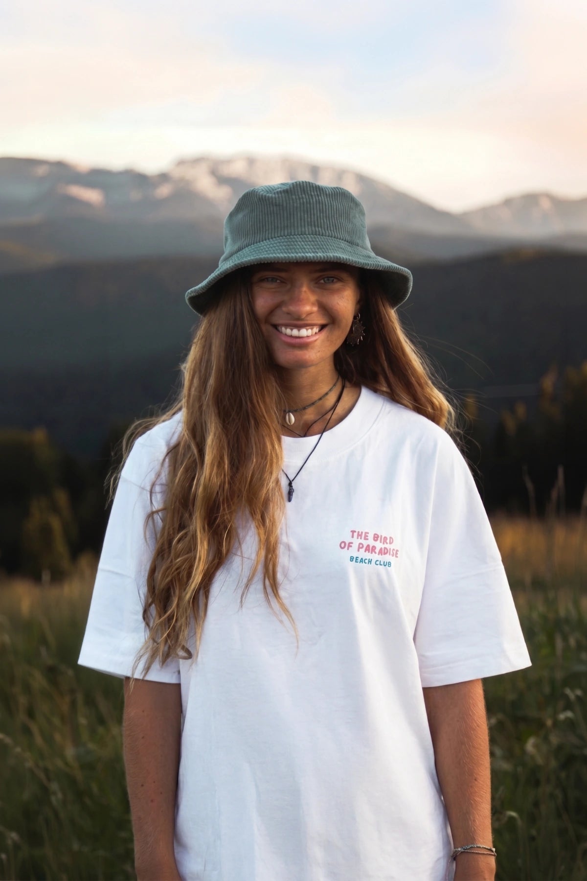 Lächelnde Frau trägt weißes Bird of Paradise Boxy Oversized T Shirt vor Berglandschaft bei goldenem Abendlicht