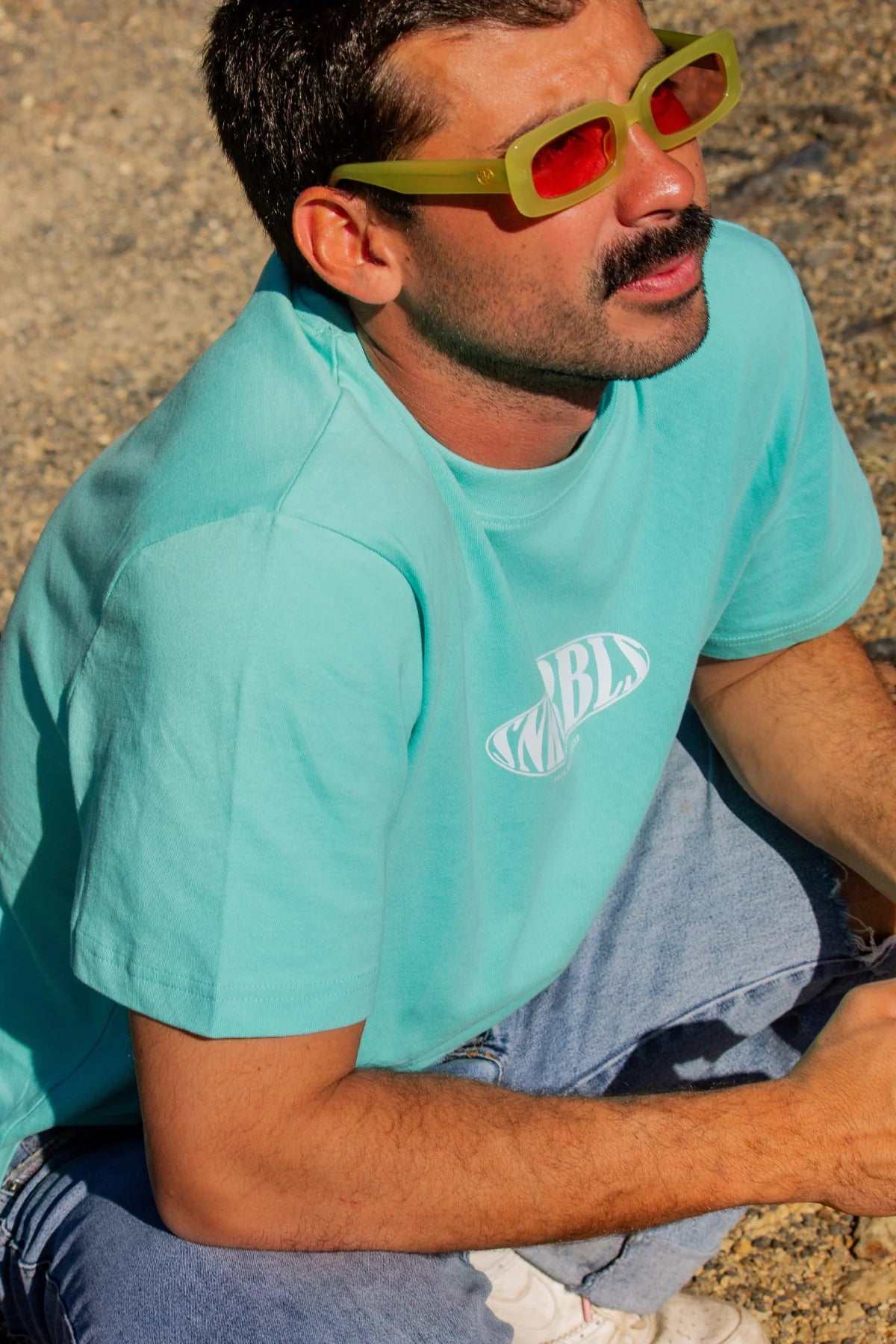 Nahaufnahme eines Mannes in türkisblauem SNKRBLS Shirt und auffälliger Sonnenbrille – relaxter Skatermoment im Sommerlicht