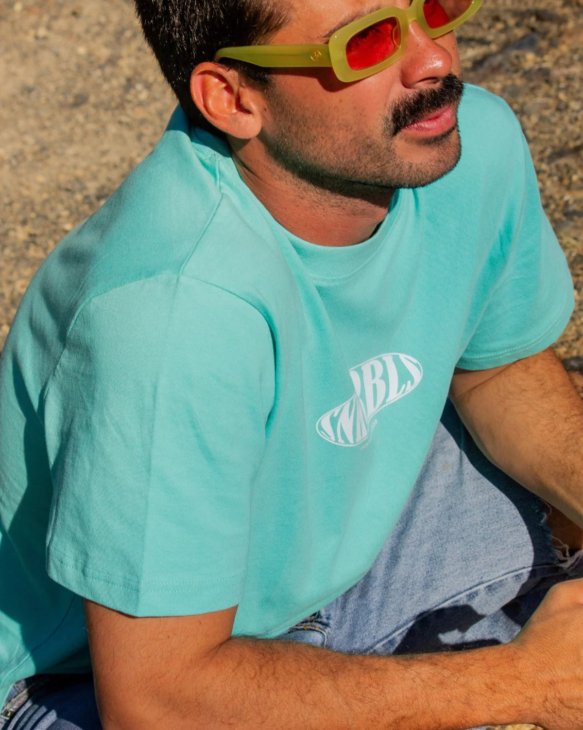 Nahaufnahme eines Mannes in türkisblauem SNKRBLS Shirt und auffälliger Sonnenbrille – relaxter Skatermoment im Sommerlicht