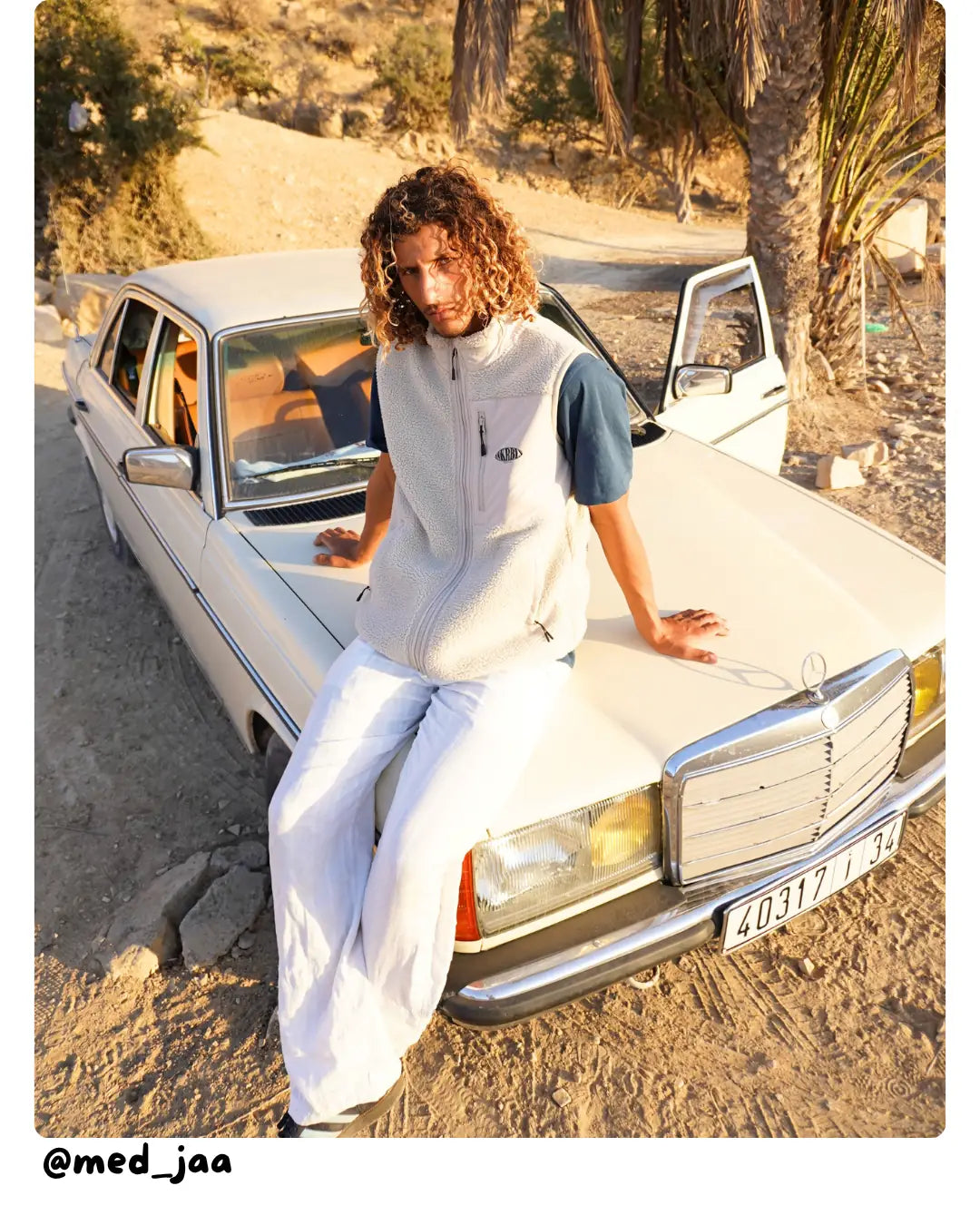 Model von The Sunkissed Rebels sitzt auf einem klassischen Mercedes in sonniger Wüstenlandschaft und trägt eine Sherpa-Weste.