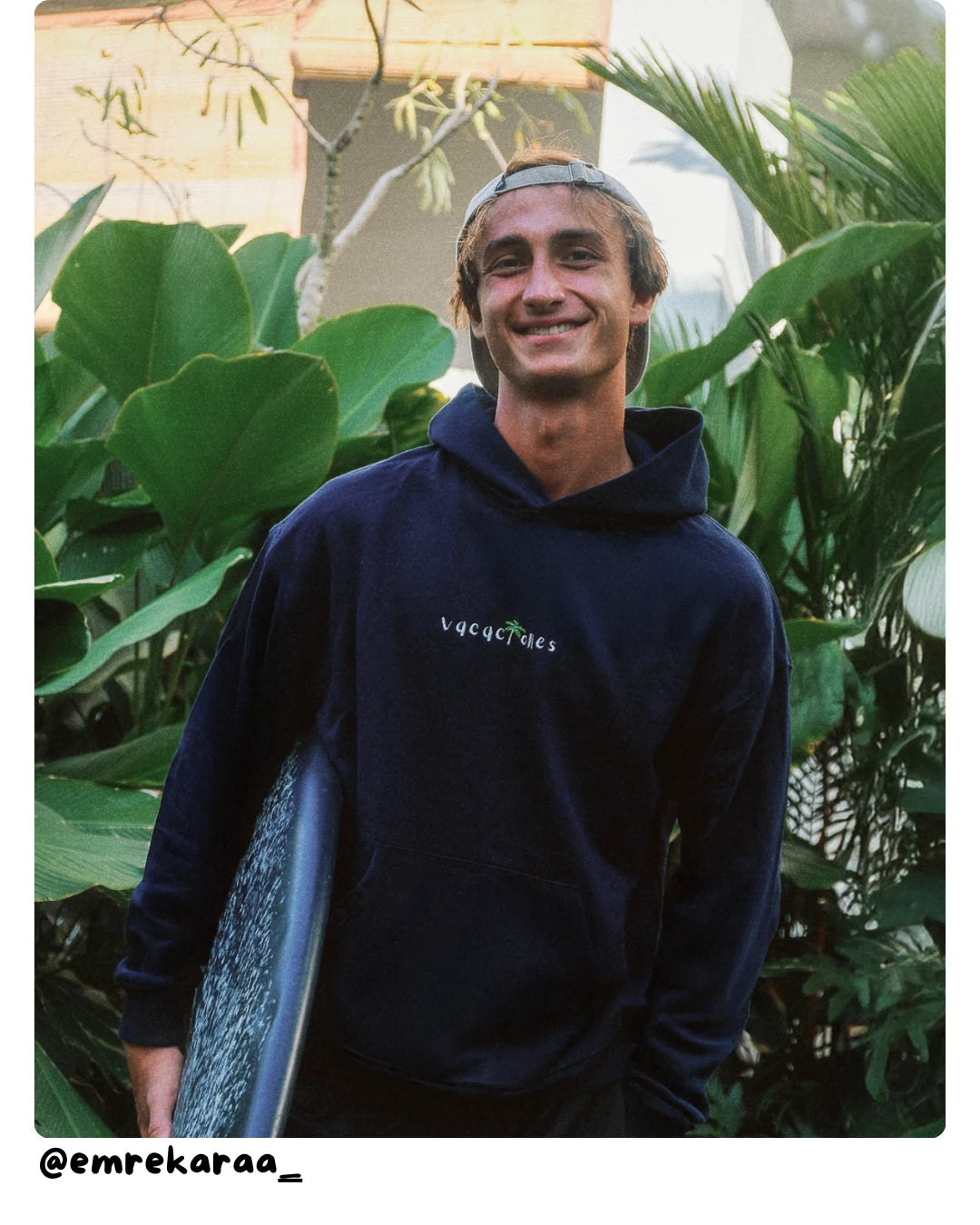 Model von The Sunkissed Rebels trägt einen dunkelblauen Surf Hoodie und hält ein Surfboard im tropischen Garten.