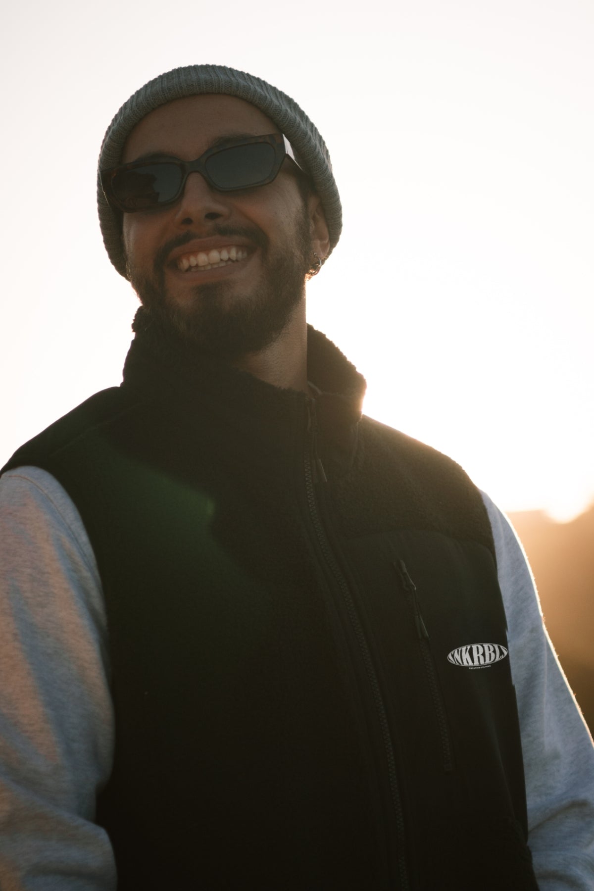Mann lacht in Kamera bei Sonnenuntergang und trägt schwarze outdoor Weste