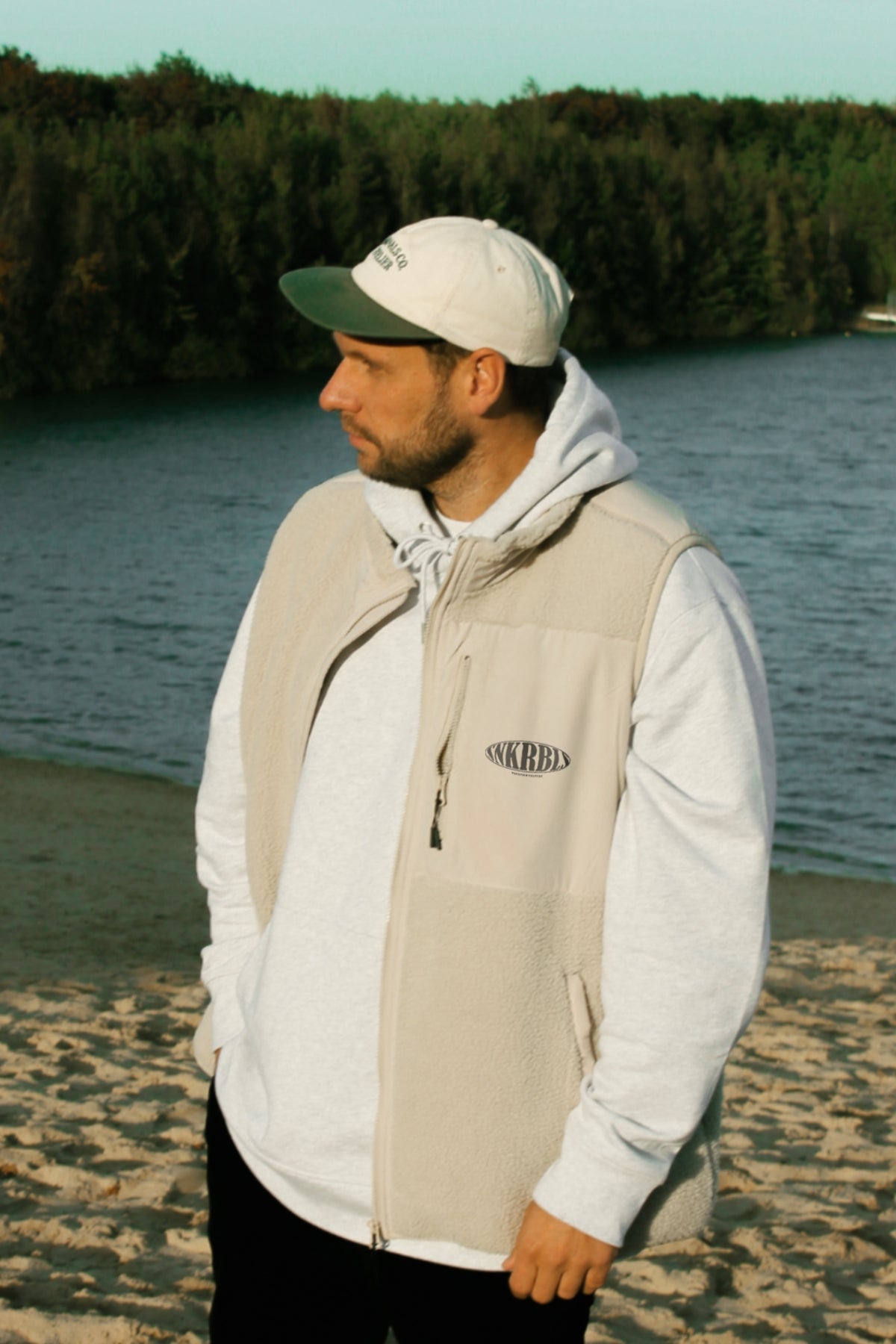 Mann steht am Strand mit Hand in der Tasche. Trägt eine Fleece Weste und Kappe