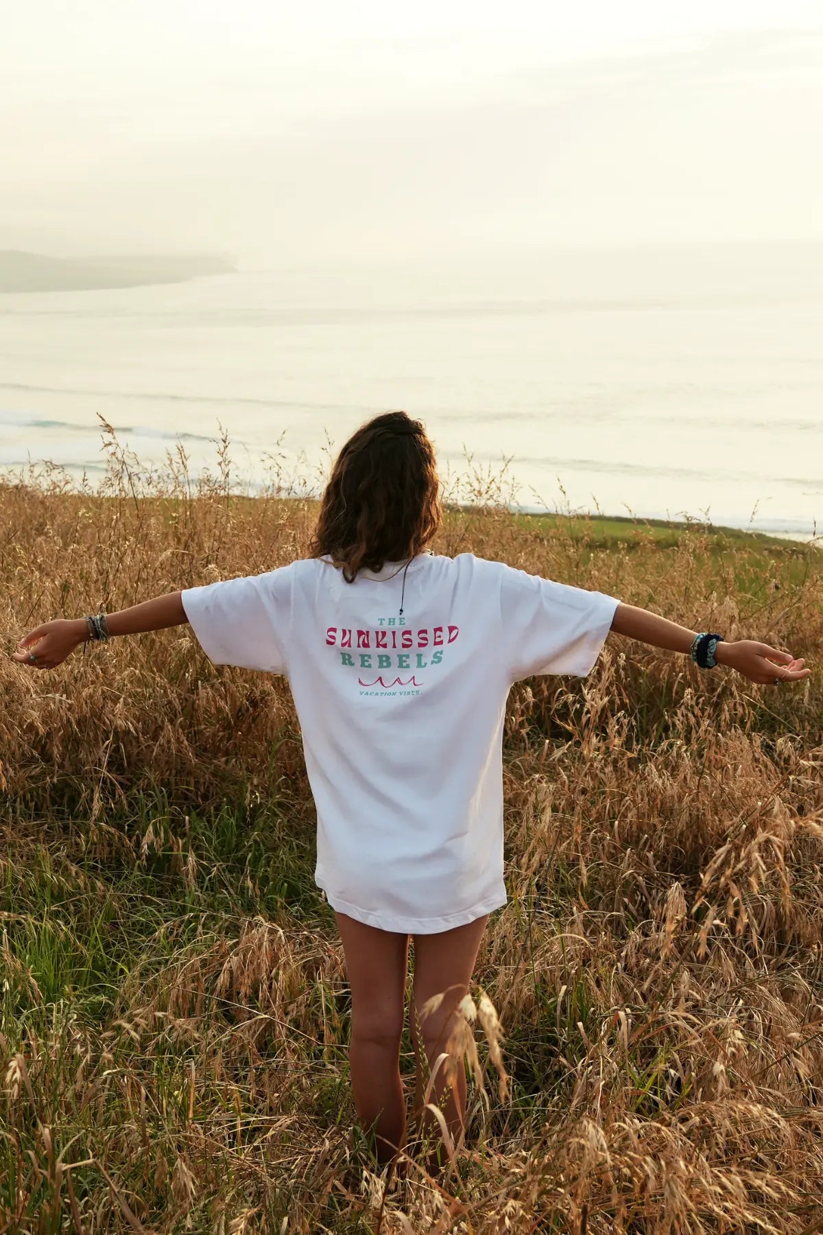 Freiheitsmoment am Meer – Frau mit ausgestreckten Armen im Vacation Vibes Shirt im Sonnenuntergang