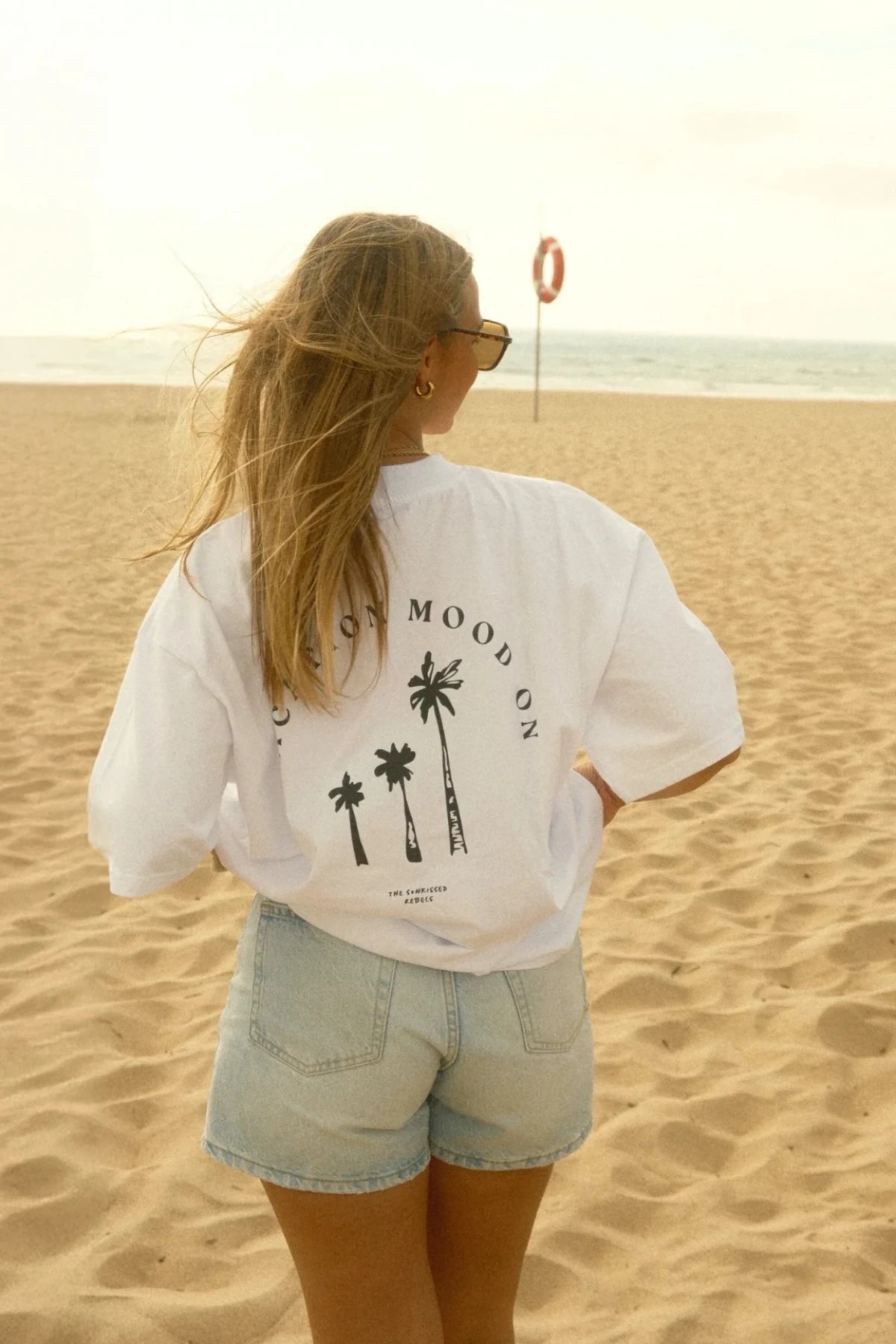Frau mit Sonnenbrille trägt das Vacation Mood On Shirt bei goldenem Licht am Strand – Wind im Haar, Palmenprint im Fokus, Urlaubsfeeling pur
