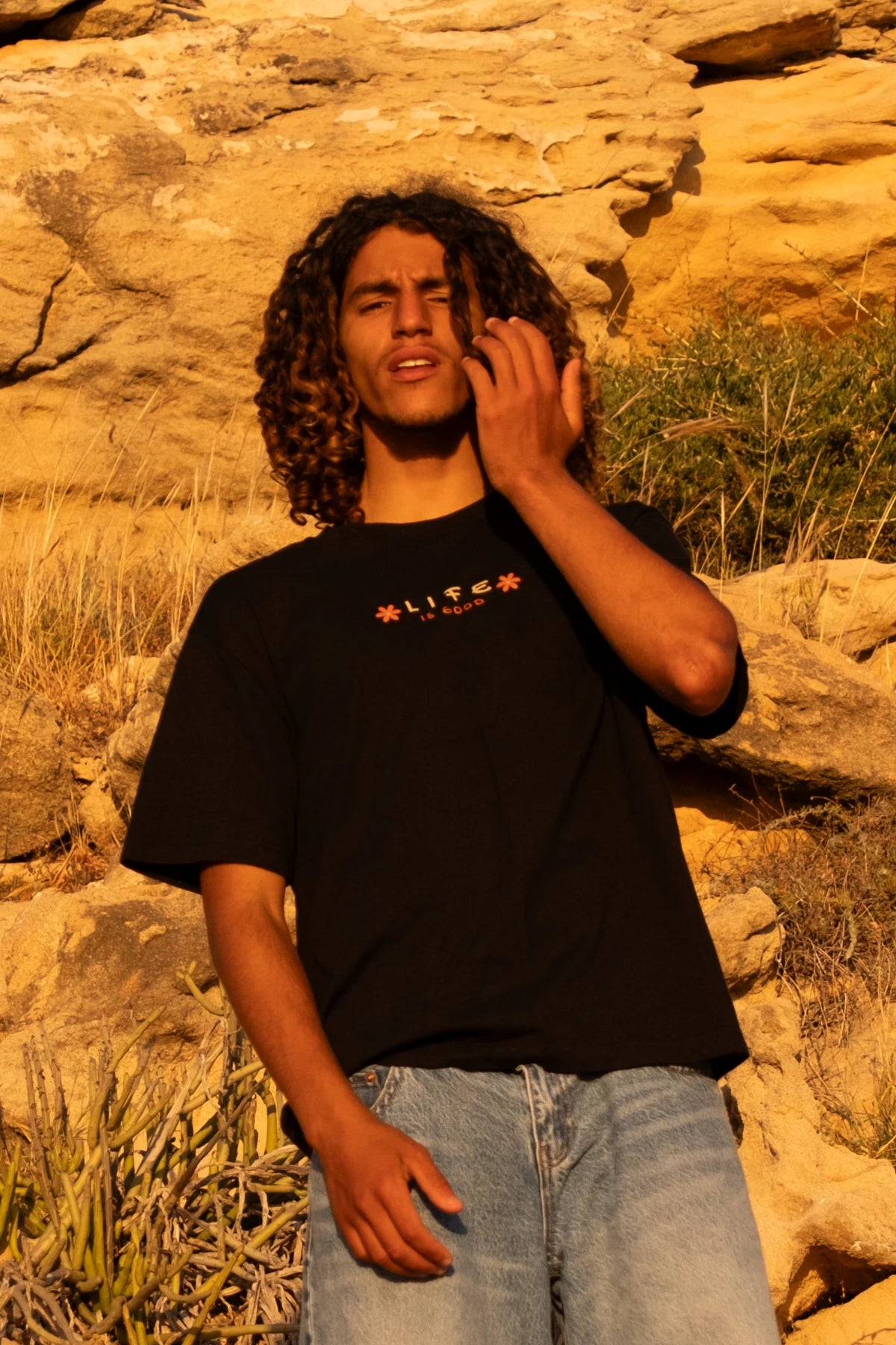 Männliches Model trägt das schwarze Good Life Light Tee vor einer felsigen Küstenlandschaft im warmen Abendlicht mit entspanntem Surf- und Lifestyle-Look.