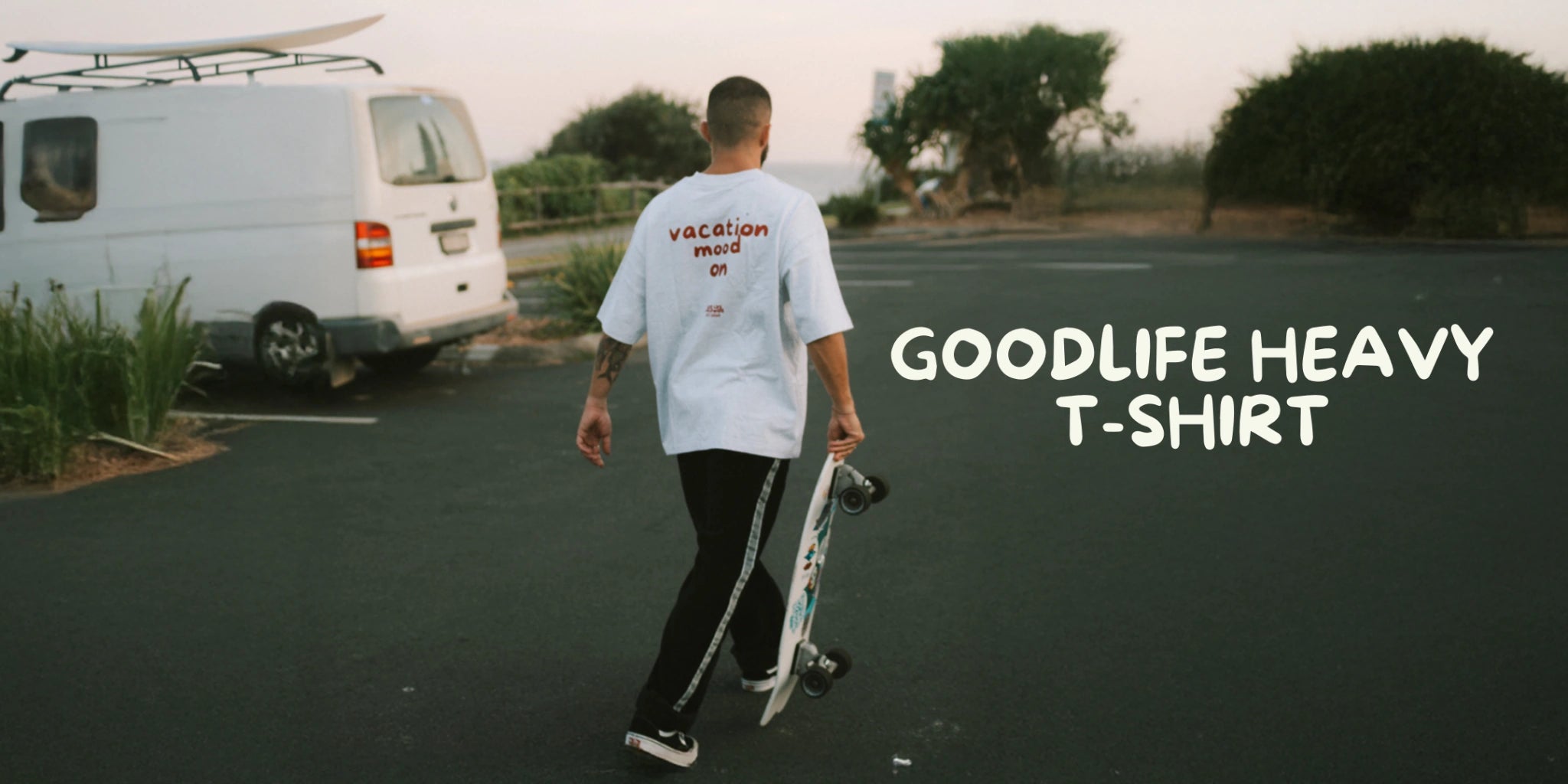 Person mit Skateboard auf Parkplatz neben Camper, trägt weißes Goodlife Heavy T-Shirt mit Rückenprint – Vacation Mood Surf Look
