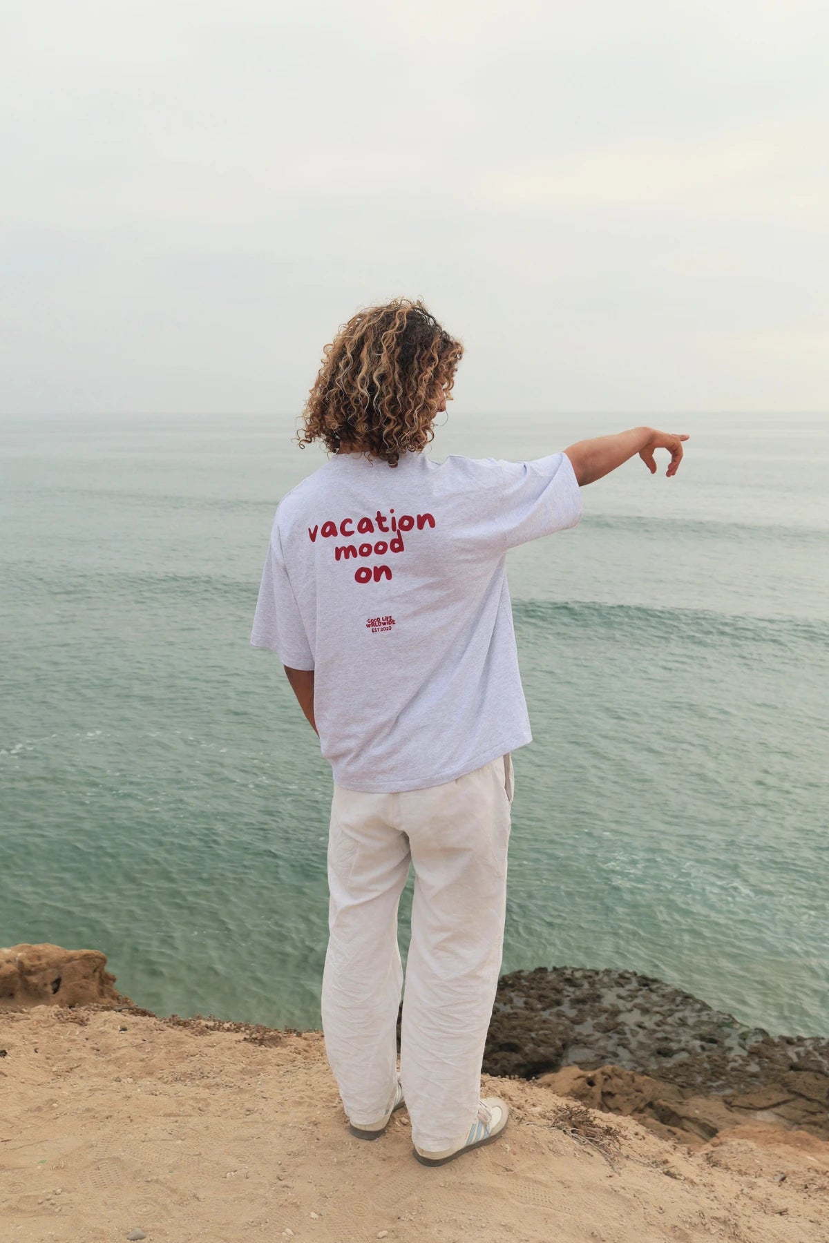 Model von The Sunkissed Rebel steht am Rand einer Klippe mit Blick aufs Meer und trägt das Goodlife T-Shirt in Cool Grey mit Rückenprint.