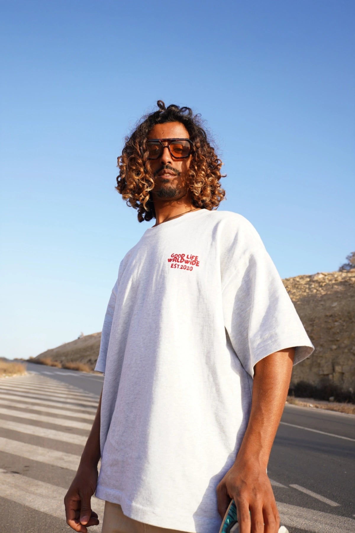 Nahaufnahme eines Models von The Sunkissed Rebel in der Sonne auf einer leeren Straße, trägt das Goodlife T-Shirt in Cool Grey und hält ein Skateboard.