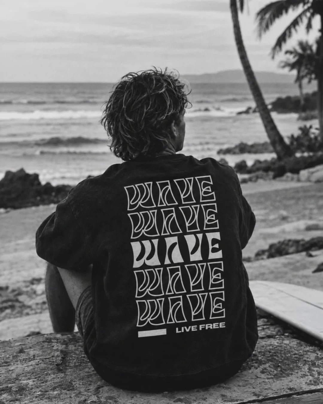 Mann sitzt am Strand und trägt vinateg schwarzen Sweater von The Sunkisse dRbeels mit Backprint Live Free
