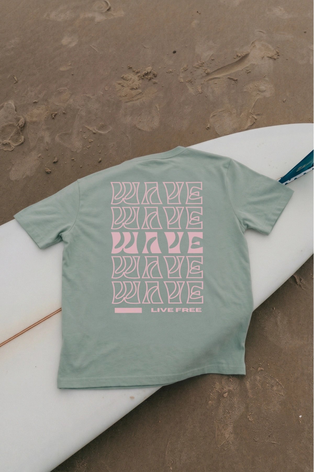 Aloegrünes Live Free Oversized T-Shirt von The Sunkissed Rebels auf Surfboard am Strand fotografiert