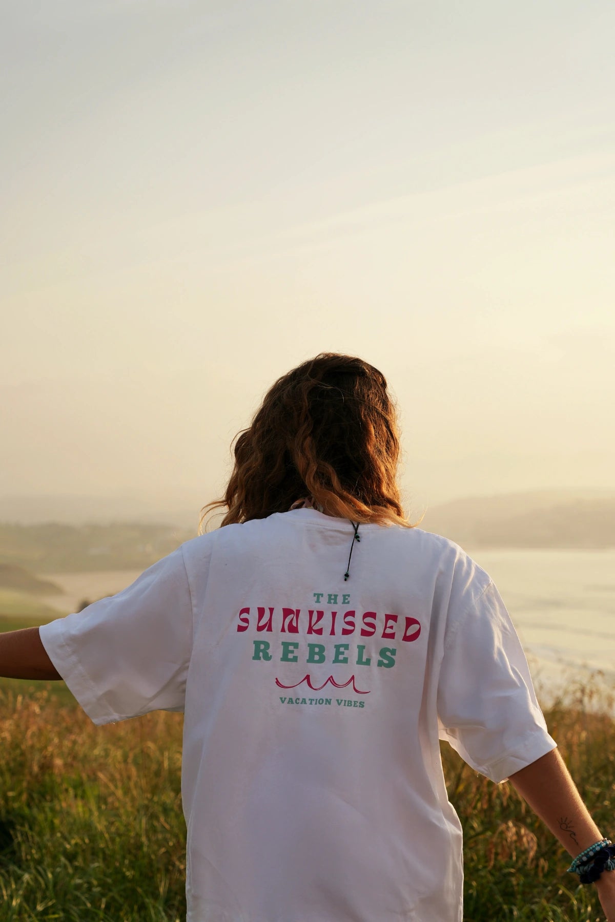 Frau mit offenen Armen im weißen Oversized T-Shirt genießt die Aussicht aufs Meer bei Sonnenaufgang