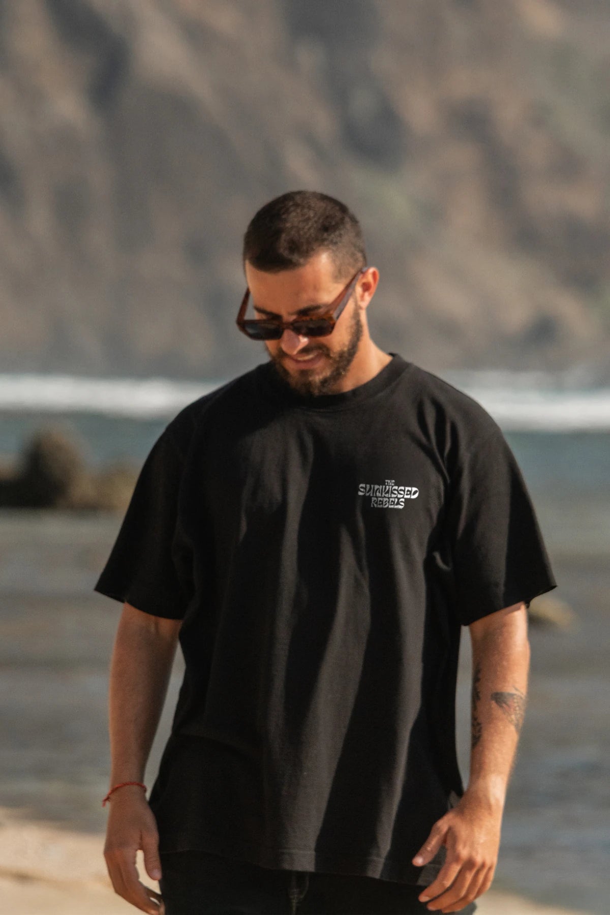 Model mit schwarzem The Sunkissed Rebels Oversized T-Shirt mit weißem Logo auf Brust am Meer