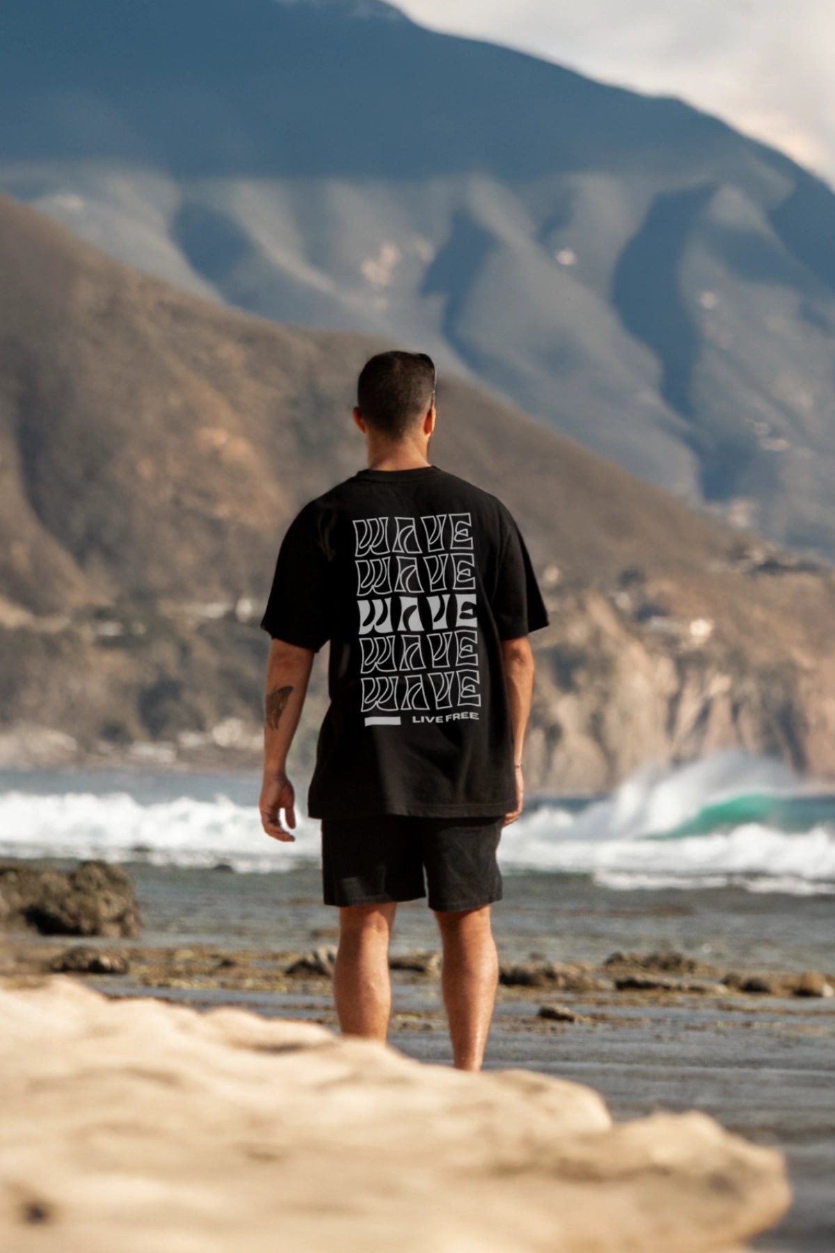 Model blickt aufs Meer in schwarzem Oversized T Shirt mit Wellen motiv mit Bergkulisse – The Sunkissed Rebels