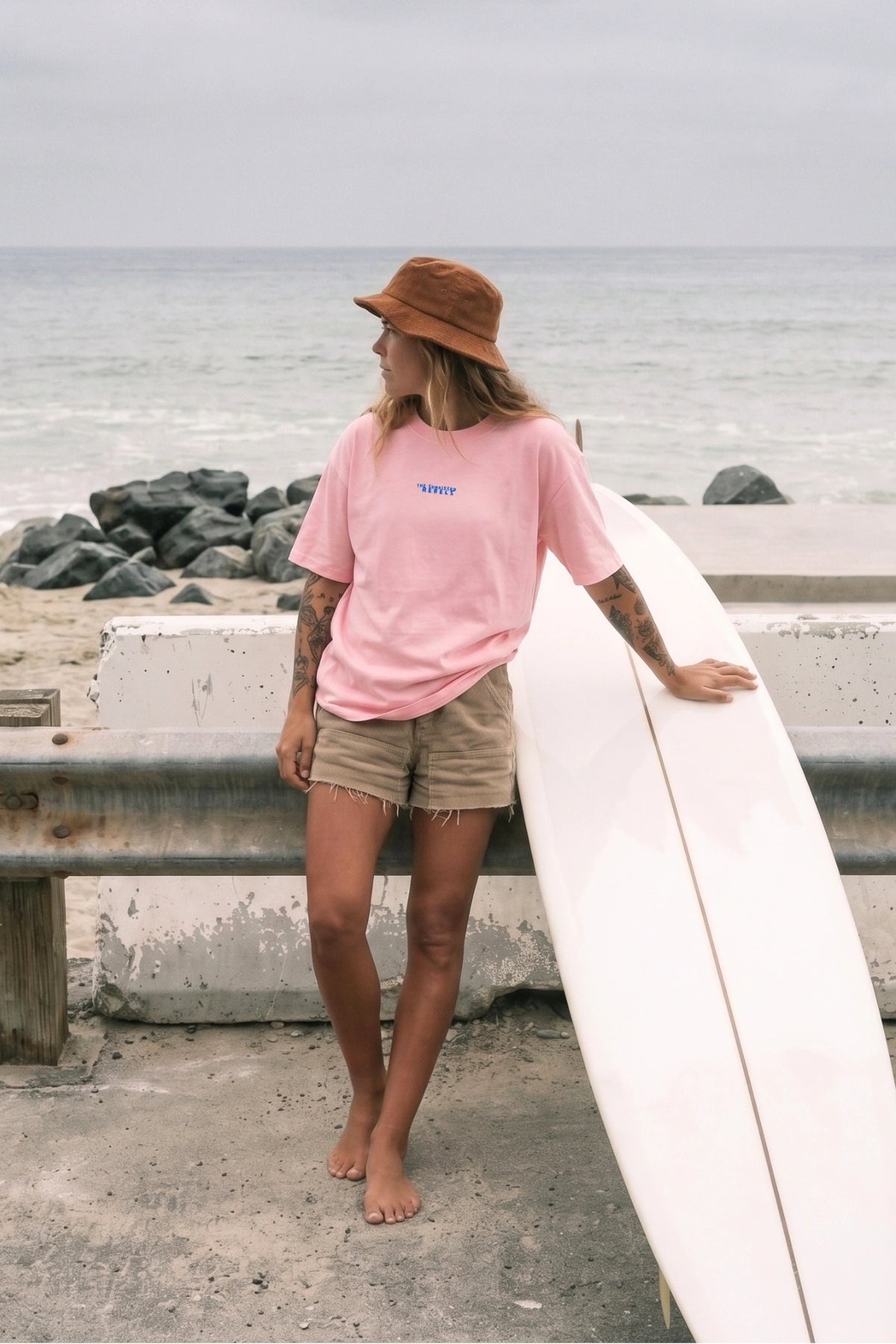 Frau trägt das No Bullshit Whale Boxy Oversized T-Shirt in Pink Joy mit Surfboard am Strand in entspannter Surf-Atmosphäre.