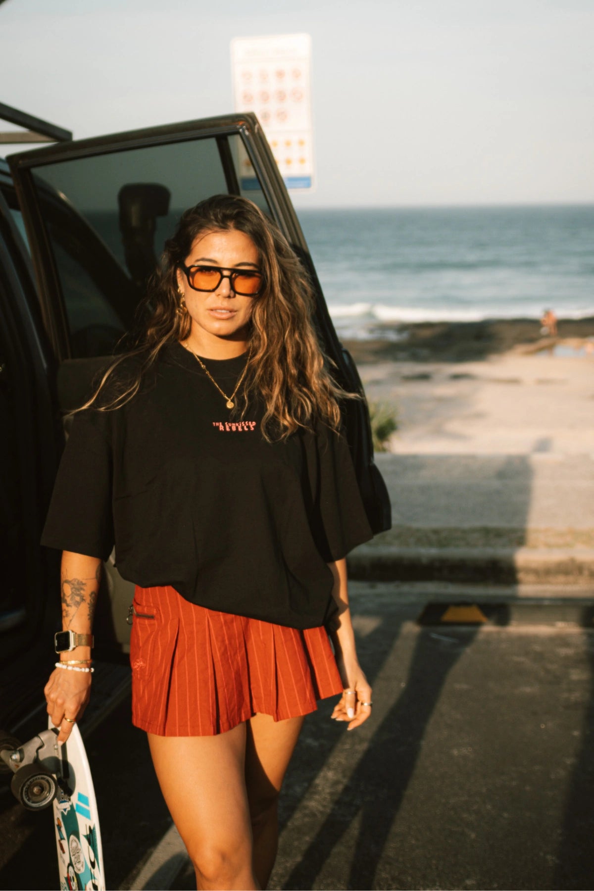 Lächelnde Frau trägt das No Bullshit Whale T-Shirt in Schwarz und roten Minirock vor dem Strand.