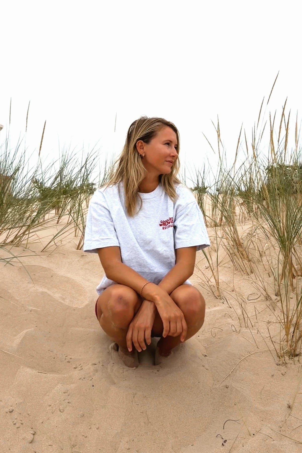 Frau sitzt in den Dünen am Strand und trägt das Goodlife Heavy T-Shirt in Cool Grey mit kleinem Brustprint.
