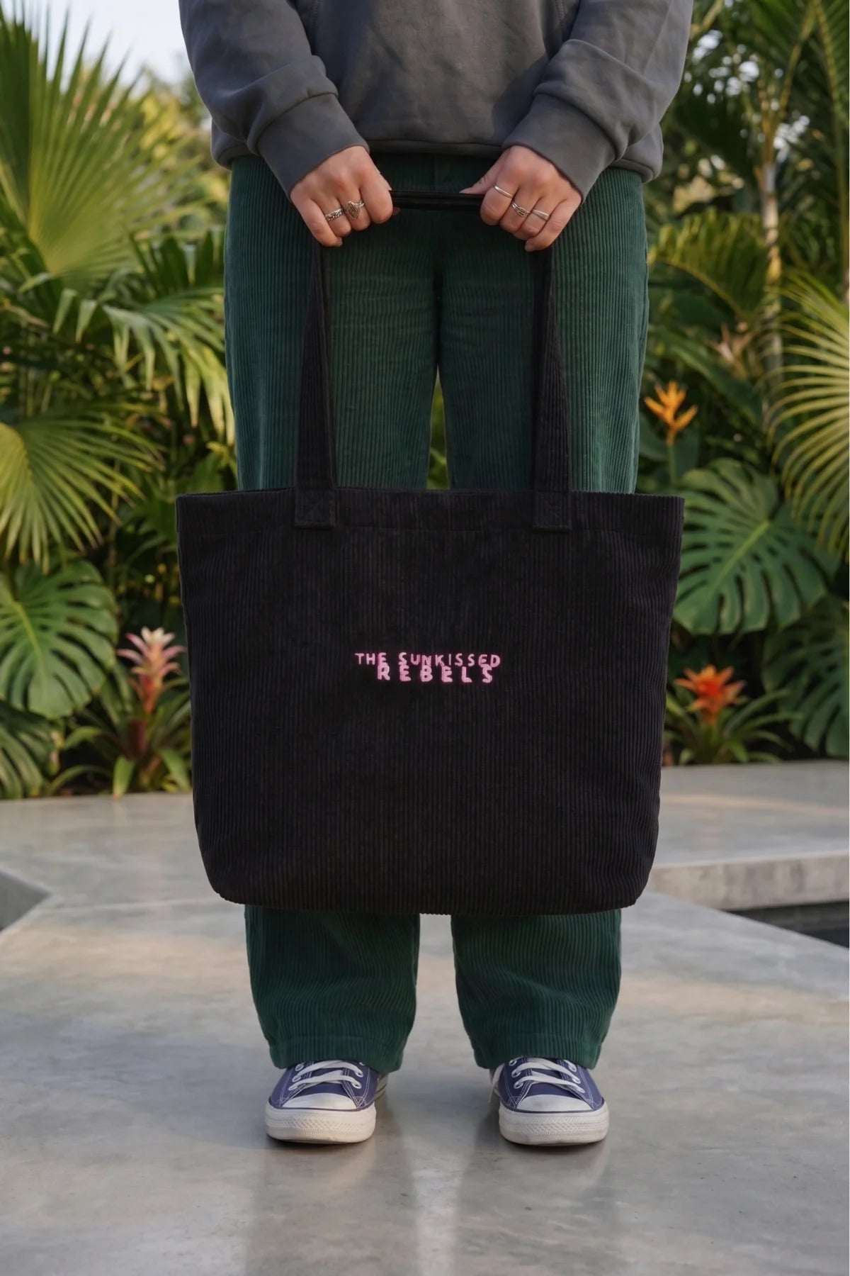 Person trägt die schwarze Cord Bag aus Bio-Baumwolle mit pinkem "The Sunkissed Rebels" Logo-Stick vor tropischem Hintergrund.