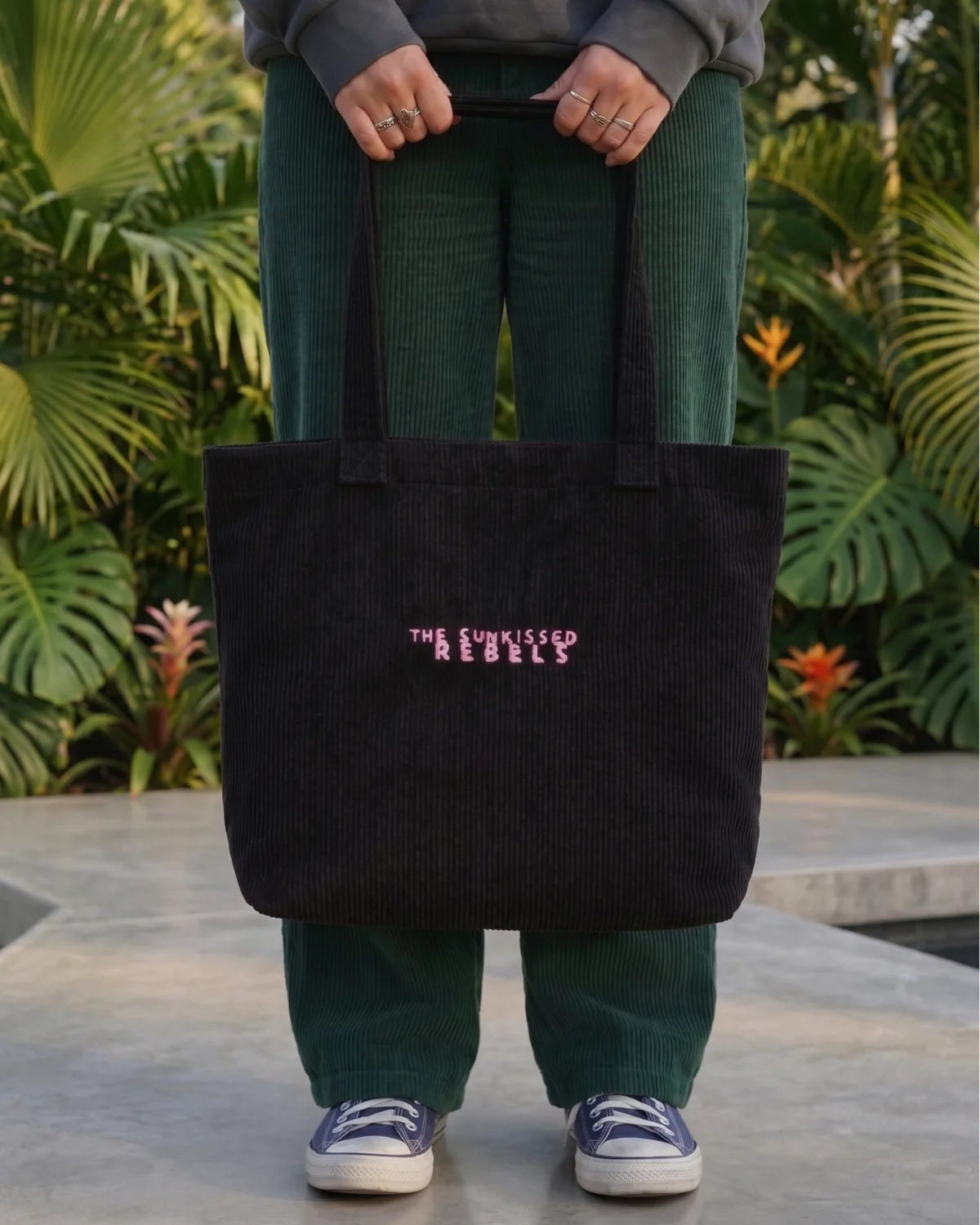 Person trägt die schwarze Cord Bag aus Bio-Baumwolle mit pinkem "The Sunkissed Rebels" Logo-Stick vor tropischem Hintergrund.