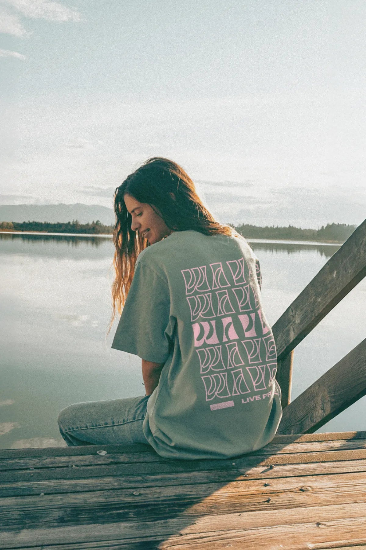 Frau im grünen Oversize-T-Shirt sitzt entspannt auf Holzsteg am See