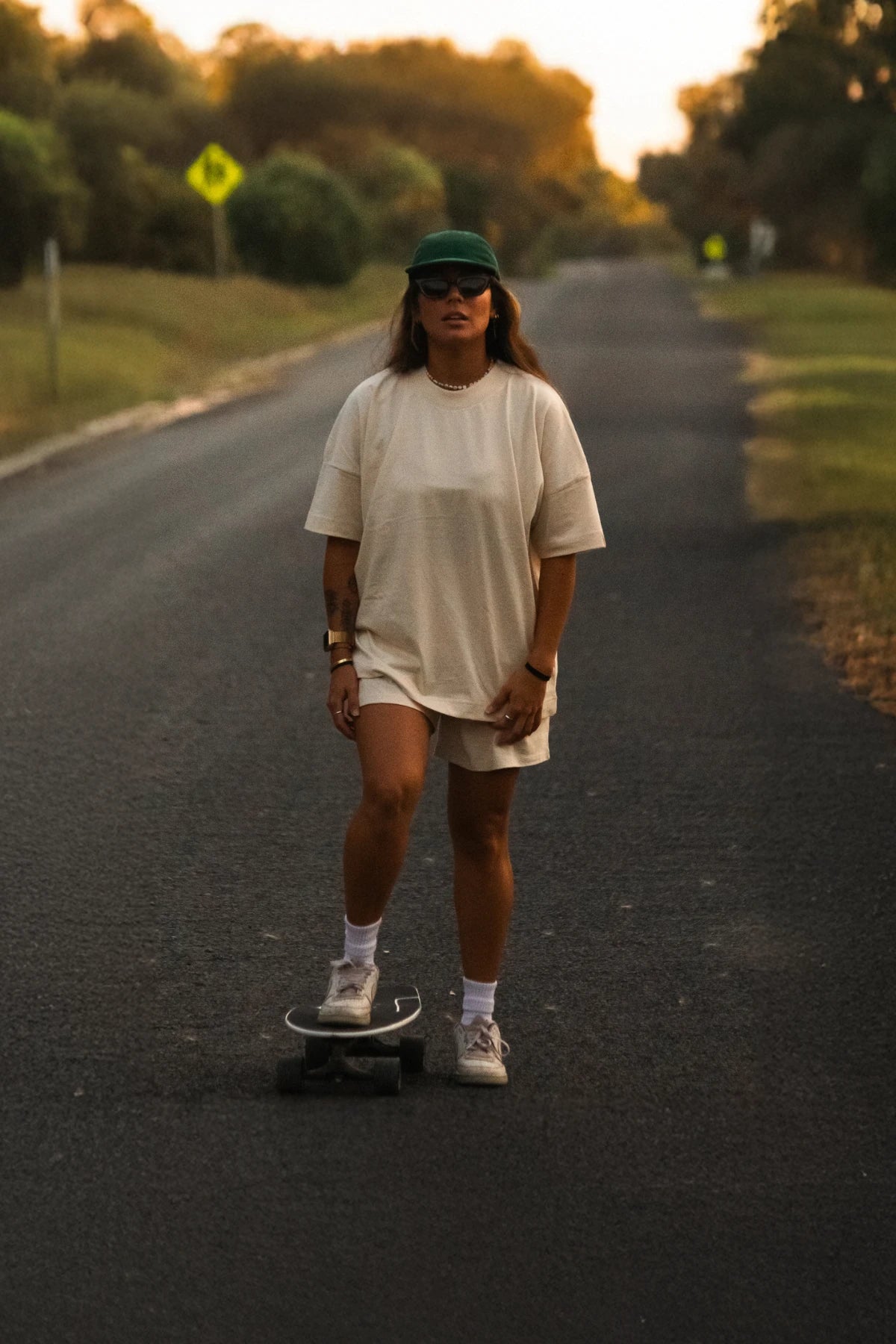Frau im naturfarbenen Oversize-T-Shirt mit Longboard bei Sonnenuntergang