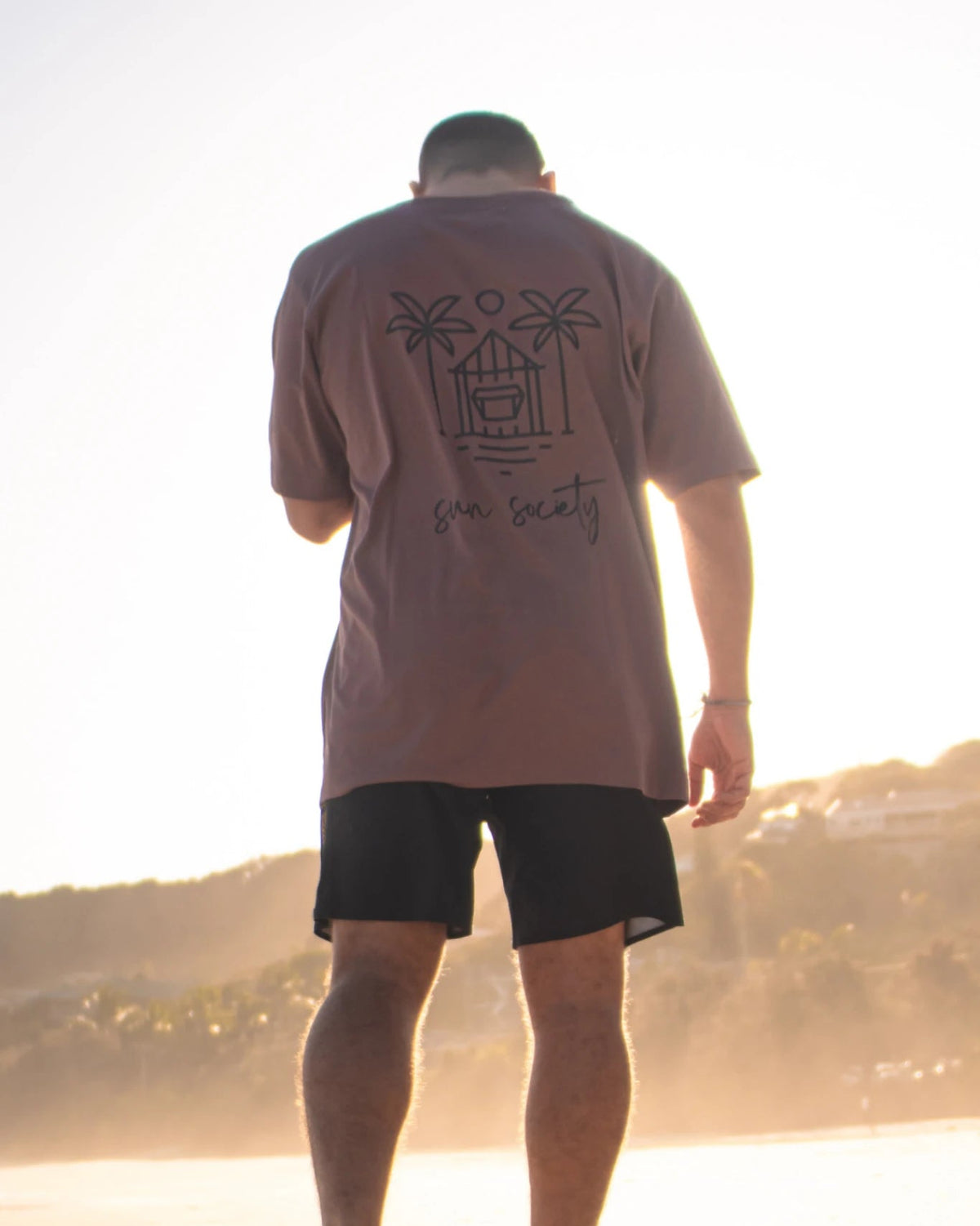 Rückenansicht des Oversized T-Shirt Açaí Sun Society von The Sunkissed Rebels am Strand bei Sonnenlicht