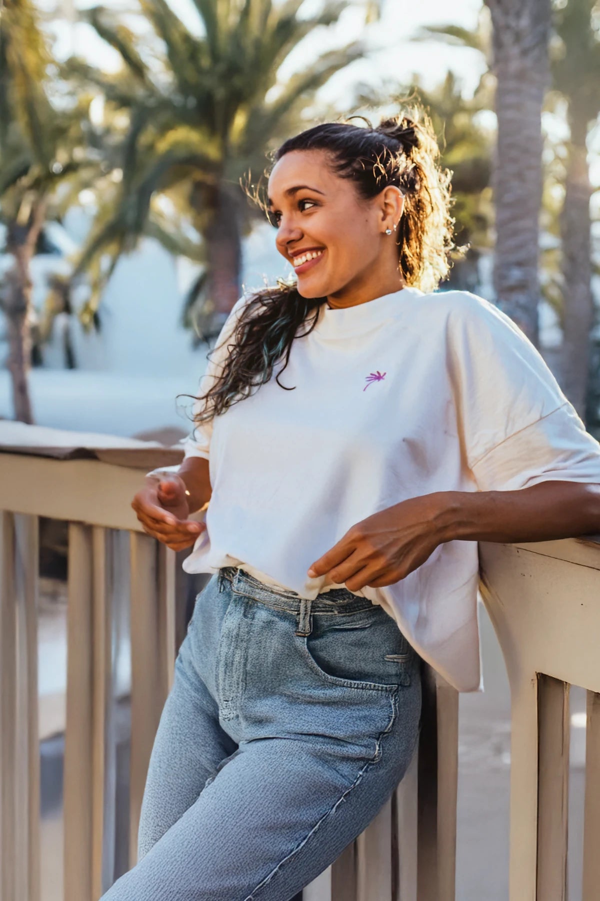 Frau mit Oversized T-Shirt in Natural Raw und pinker Palmenstickerei lehnt entspannt am Balkon.