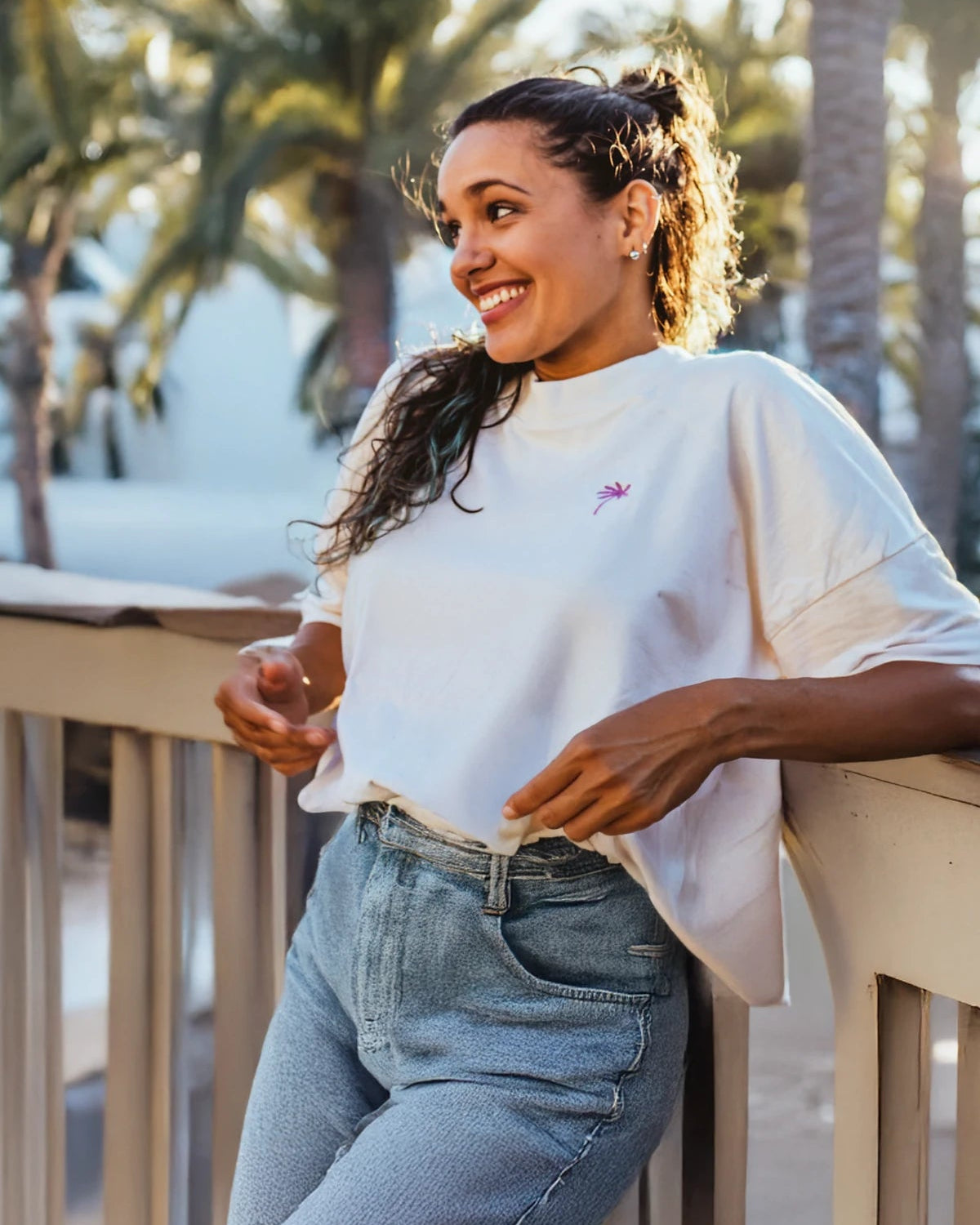 Frau mit Oversized T-Shirt in Natural Raw und pinker Palmenstickerei lehnt entspannt am Balkon.