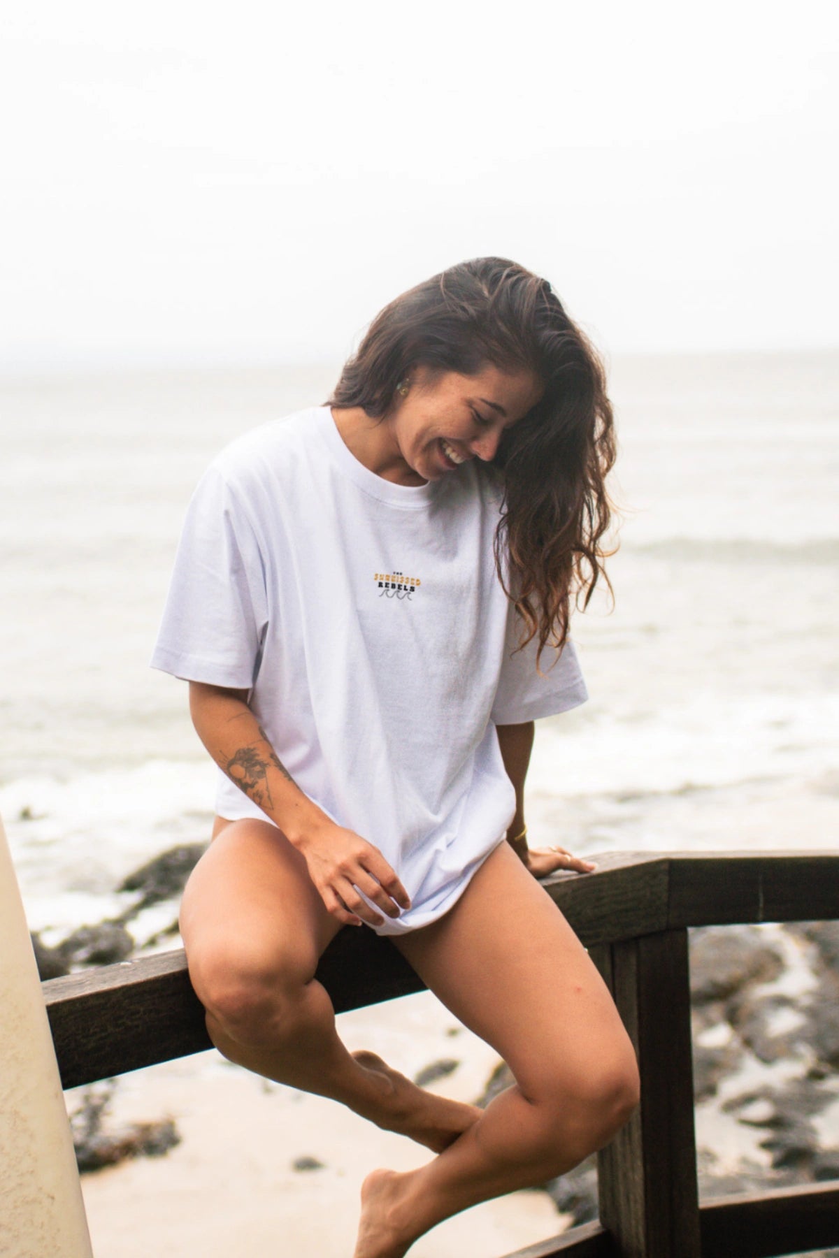 Frau sitzt auf Geländer und lacht. Sie trägt das weiße Oversized T-Shirt Paradise & Sunset von The Sunkissed Rebels.
