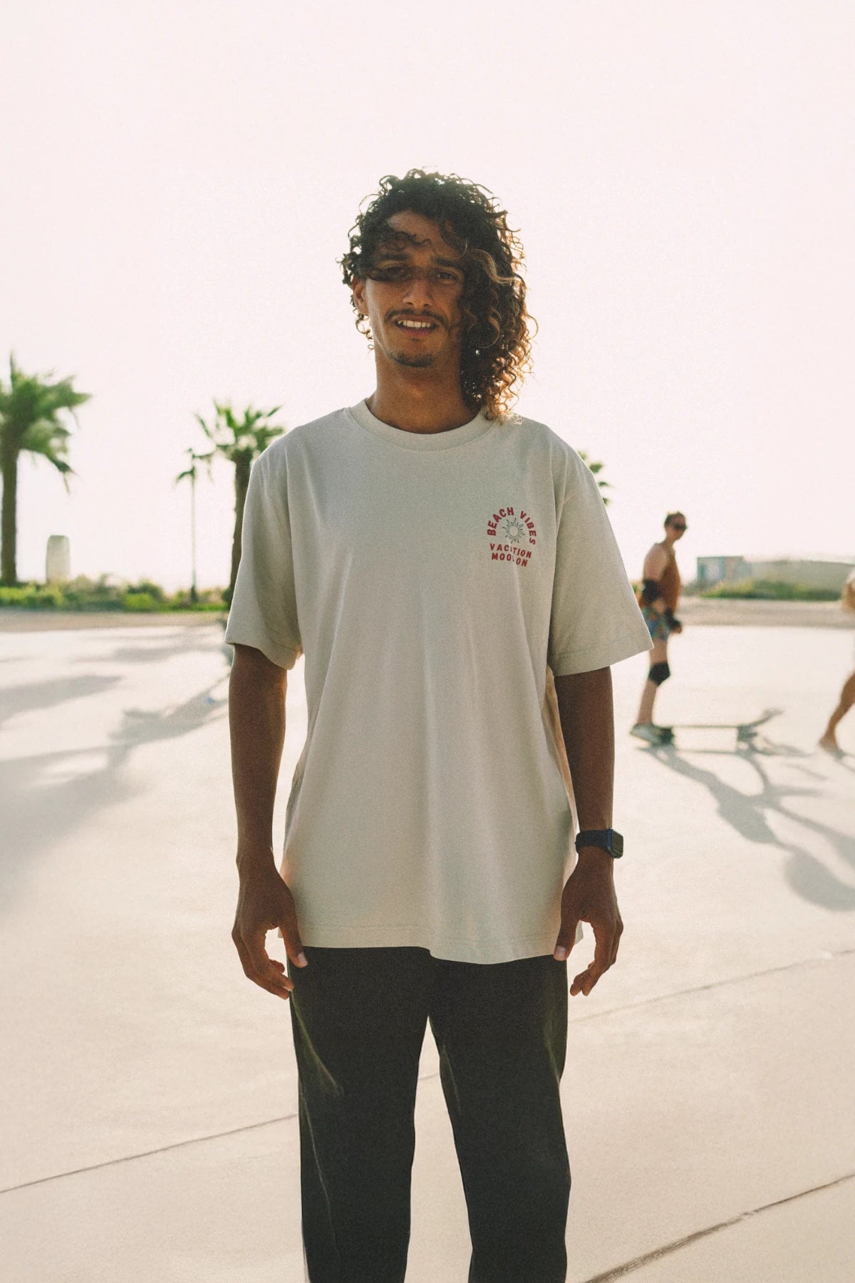 Mann trägt "Beach Vibes" Oversized T-Shirt in Sandfarbe im Sonnenlicht mit Skateboarder im Hintergrund