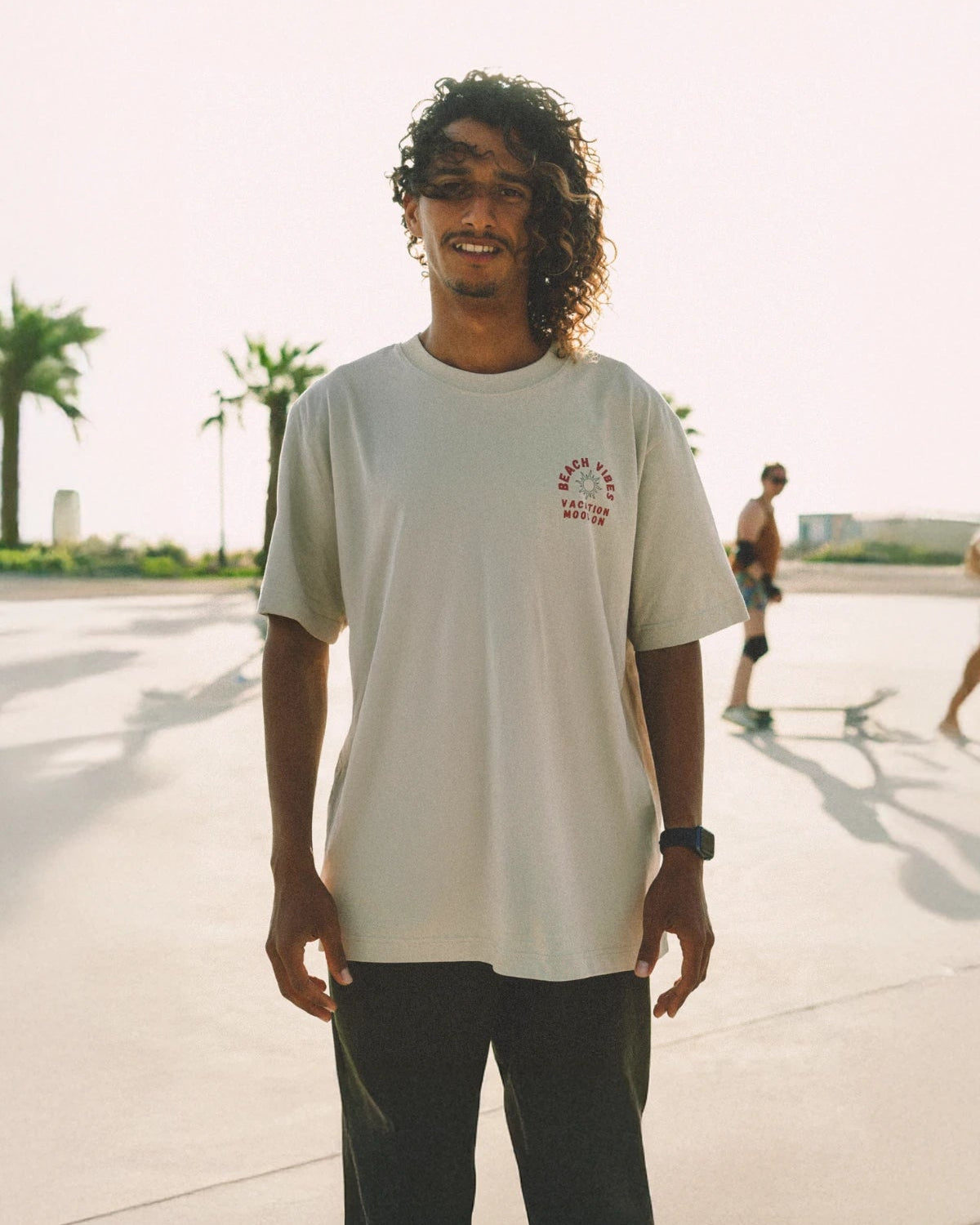 Mann trägt "Beach Vibes" Oversized T-Shirt in Sandfarbe im Sonnenlicht mit Skateboarder im Hintergrund