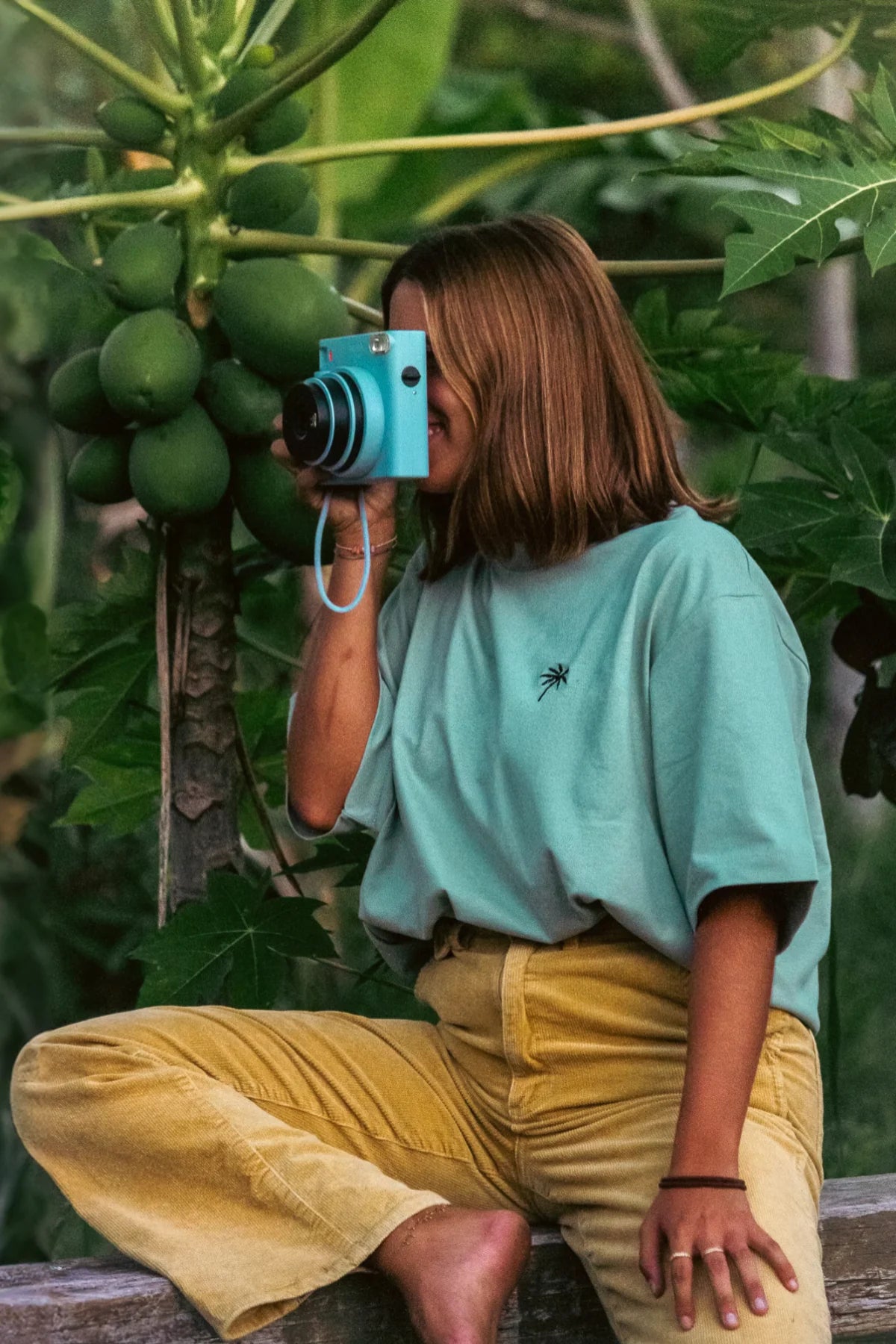 Model trägt oversized T-Shirt in aloe mit Kamera in tropischer Umgebung – The Sunkissed Rebels