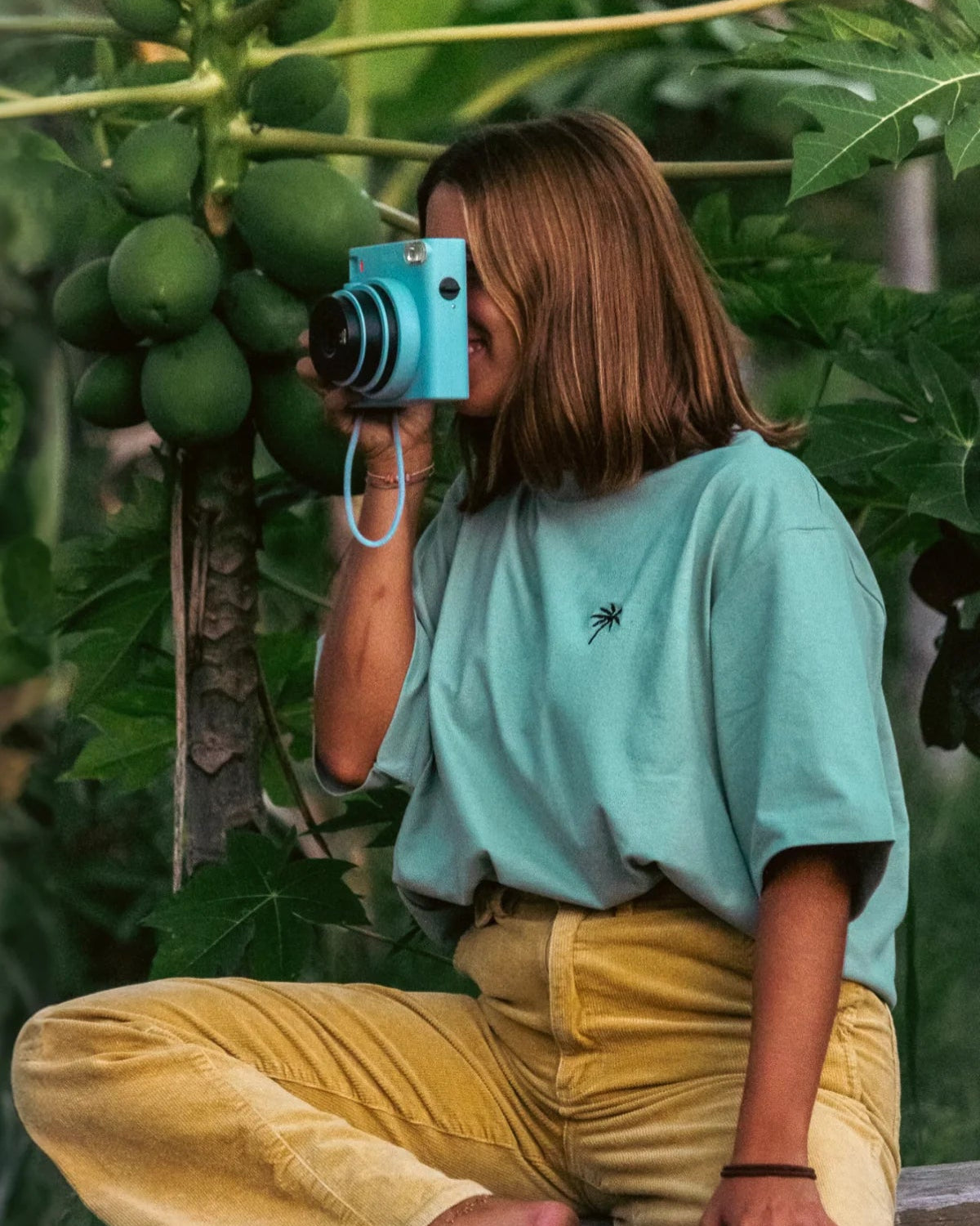 Model trägt oversized T-Shirt in aloe mit Kamera in tropischer Umgebung – The Sunkissed Rebels