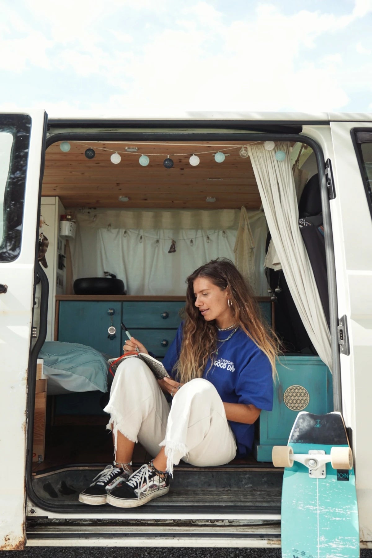 Frau sitzt in einem geöffneten Van und schreibt in ein Notizbuch, trägt ein Oversized T-Shirt mit inspirierendem Print