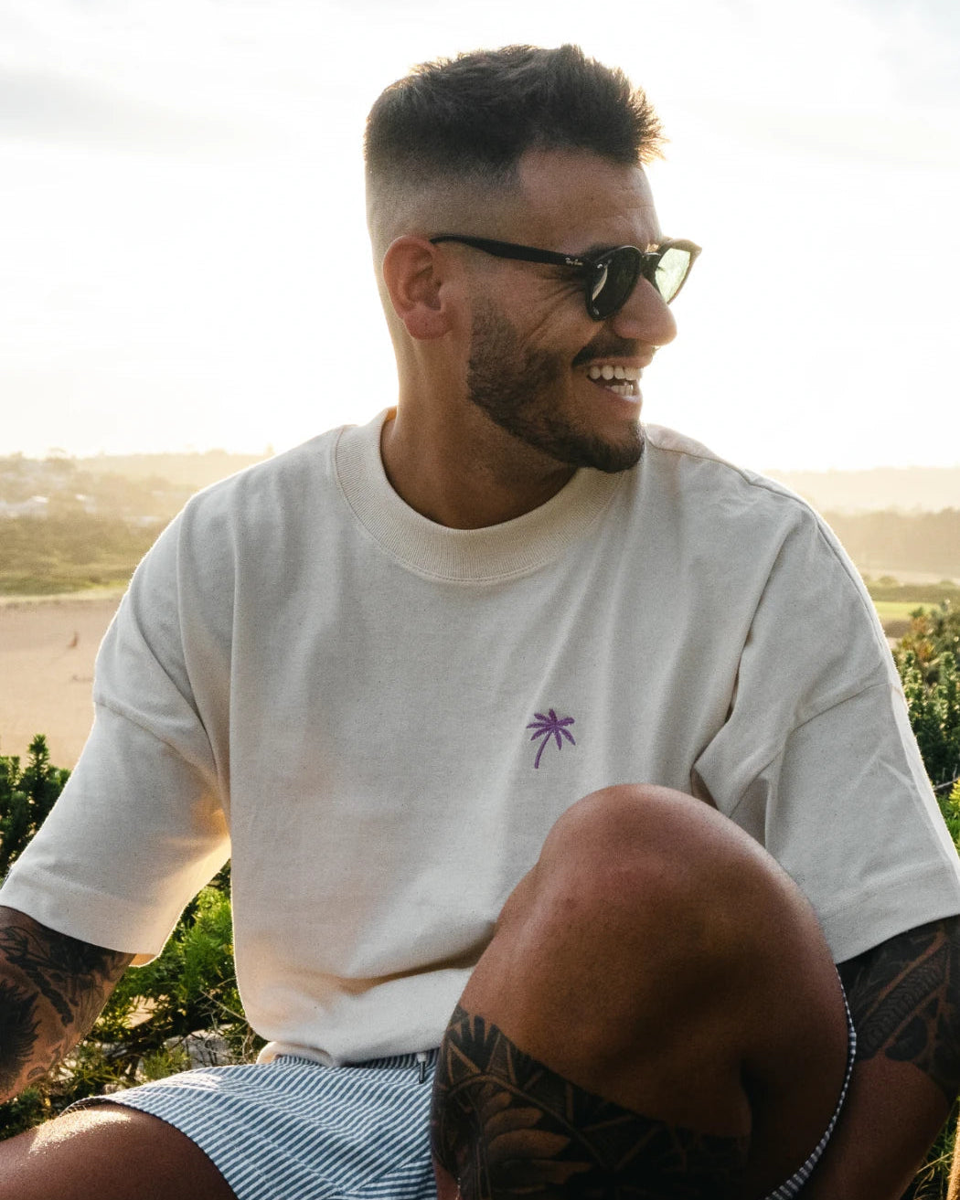Lachender Mann im oversized T-Shirt in Natural Raw mit violettem Palmen-Print, bei Sonnenuntergang in der Natur sitzend.