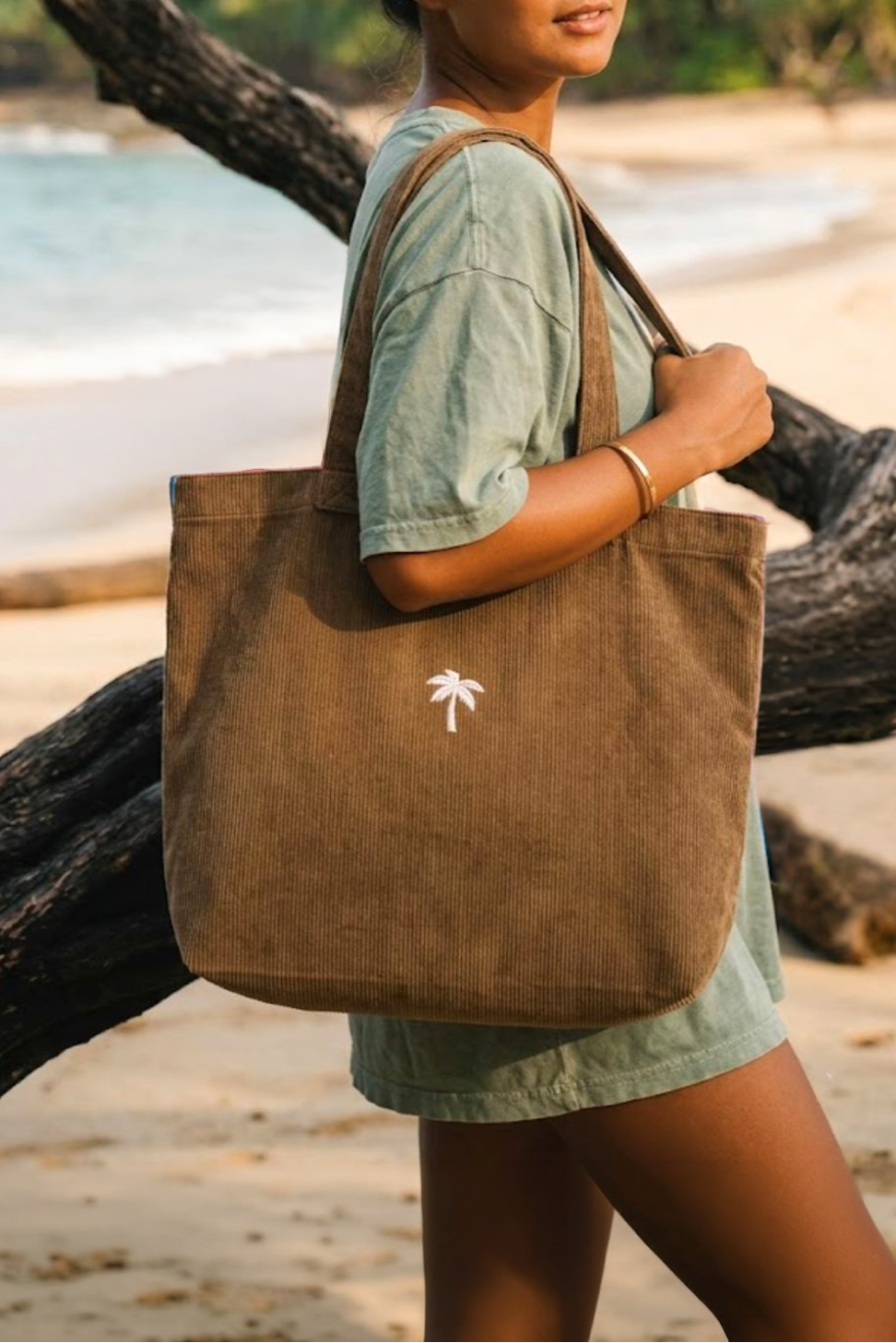 Ein Model trägt die braune Palm Tree Cord Bag aus Feincord über der Schulter an einem tropischen Strand.