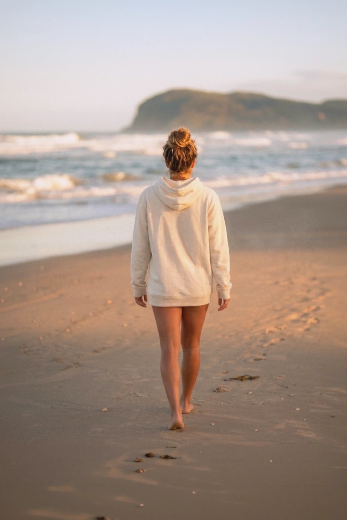 Rückansicht einer Frau läuft barfuß am Strand und trägt natürlichen Palm Tree Hoodie in Raw Beige – entspannter Beach Look bei Sonnenuntergang