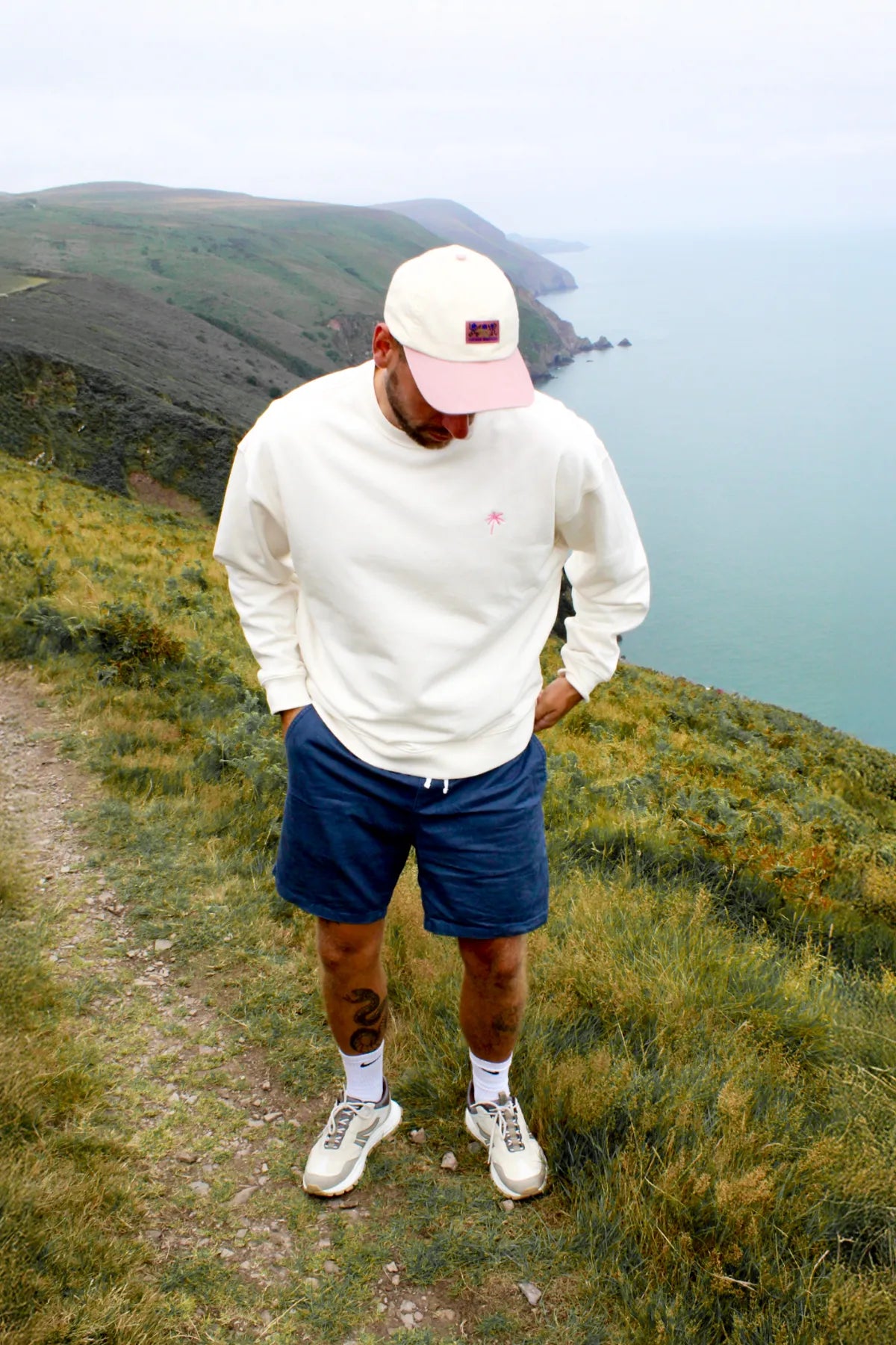 Model trägt den cremefarbenen Palm Tree Oversized Sweater bei einer Wanderung entlang eines Bergpfads mit Meerblick.
