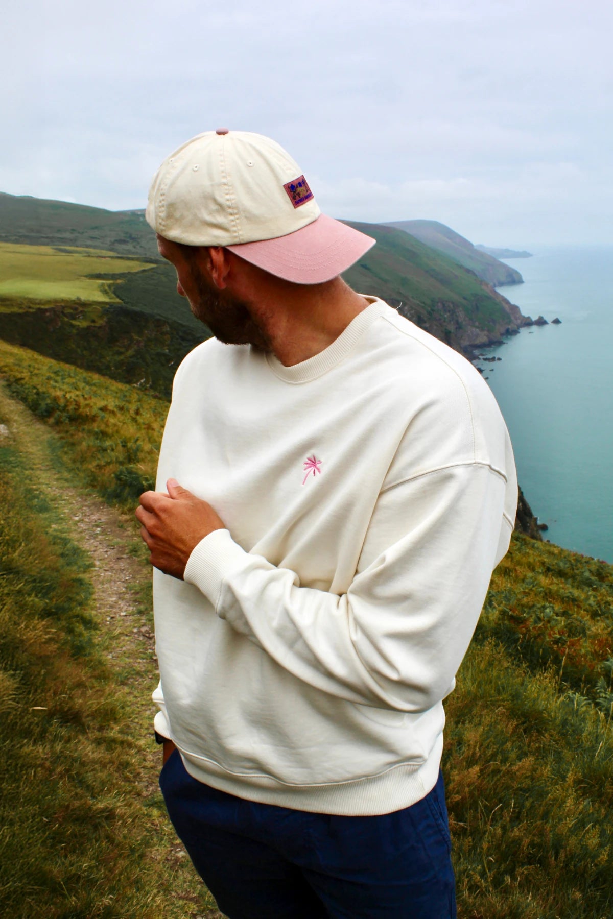 Model im cremefarbenen Sweater steht auf einem Küstenpfad und blickt aufs Meer.