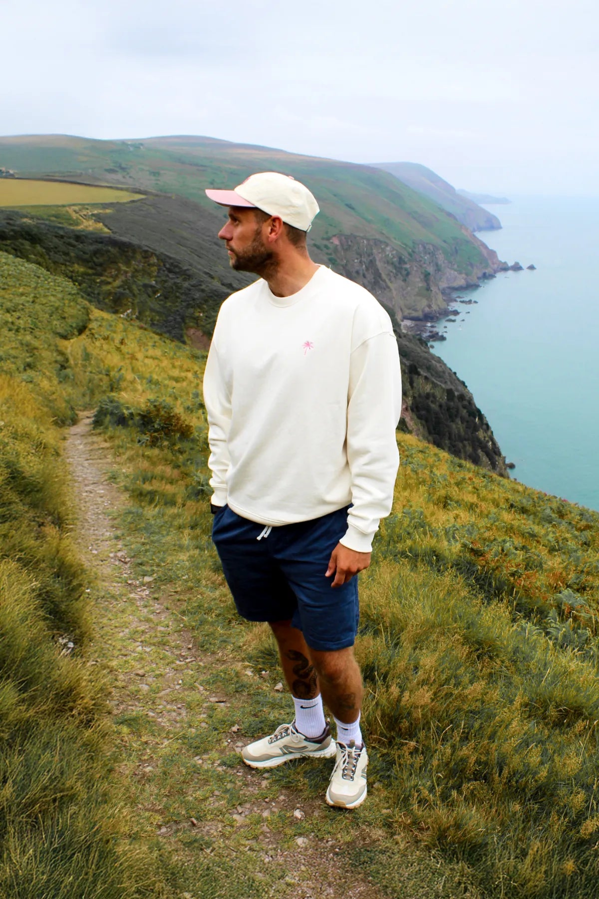 Model in cremefarbenem Oversized Sweater, seitlich zum Meer stehend, auf einem Pfad an den Klippen.