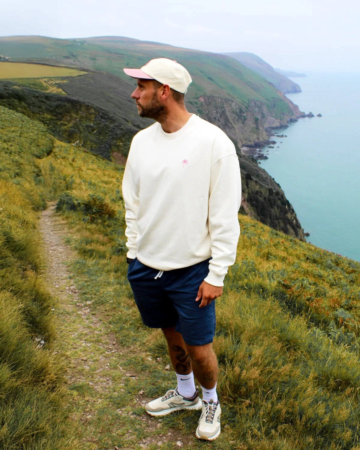 Model in cremefarbenem Oversized Sweater, seitlich zum Meer stehend, auf einem Pfad an den Klippen.