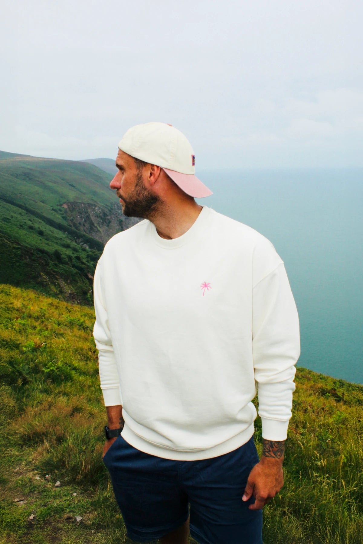 Model trägt den Palm Tree Oversized Sweater Cream in Kombination mit Shorts und Cap bei einer Wanderung an der Küste.